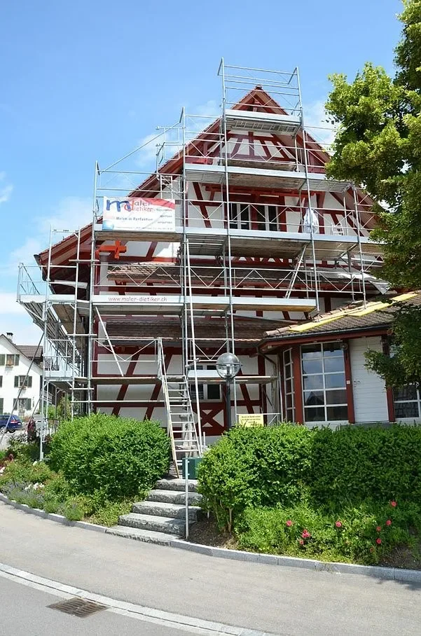 Ein Fachwerkhaus, das renoviert wird, mit Gerüsten drumherum und grünem Garten im Vordergrund.