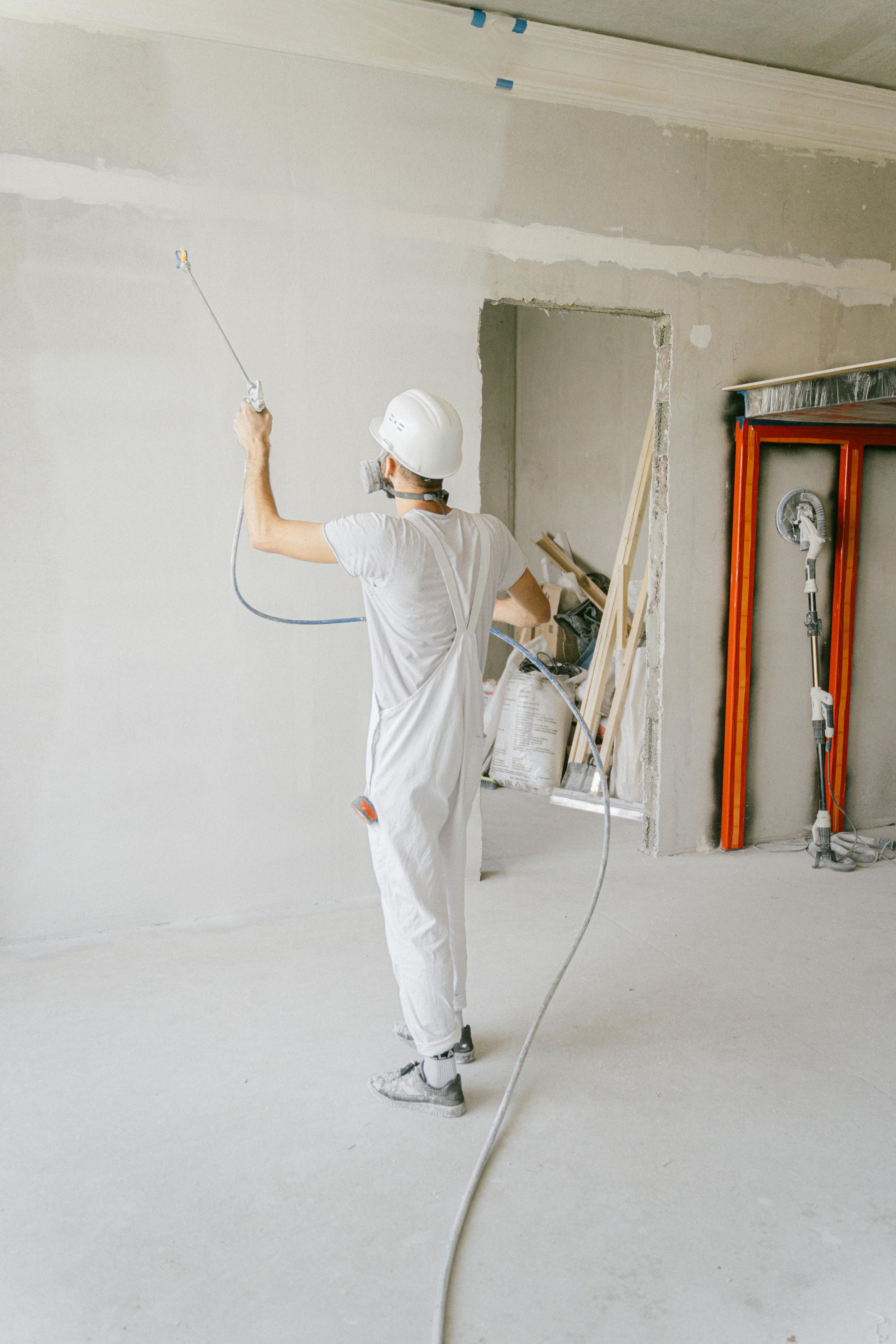 Ein Arbeiter mit Schutzhelm und Staubmaske, der eine weisse Wand streicht oder sprüht, in einem Raum unter Bauarbeiten.