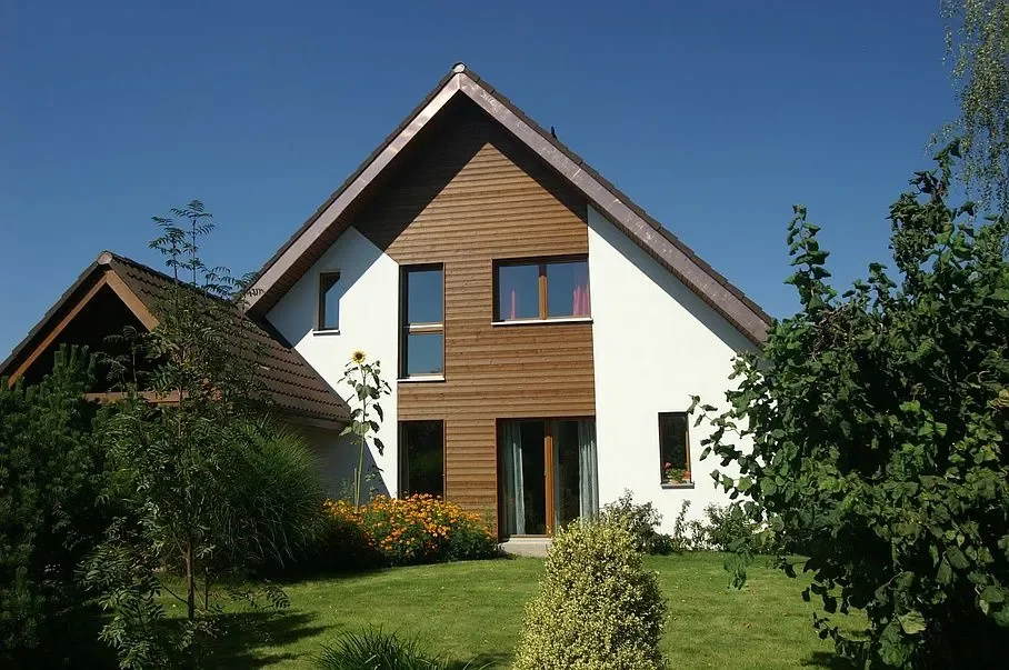 Modern zweistöckiges Haus mit Holz- und weissen Fassaden, umgeben von Garten und Bäumen, blauer Himmel.