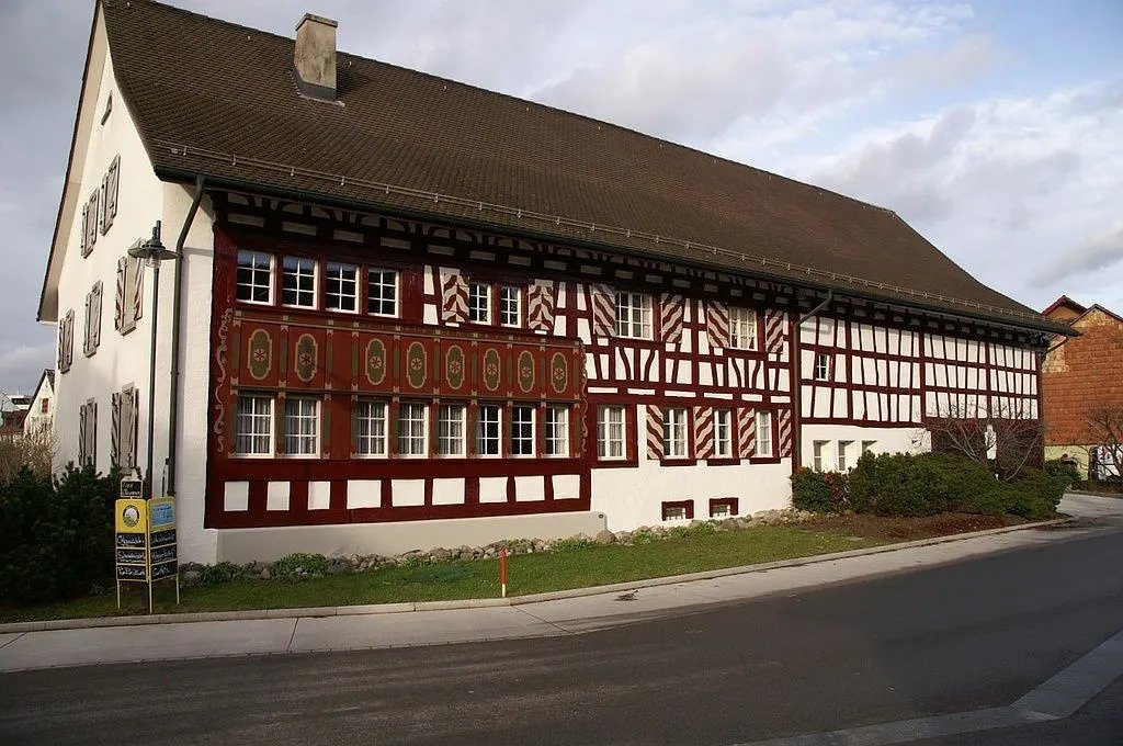 Ein Fachwerkhaus mit weissen Wänden und roten Holzstrukturen, das an einer Strasse steht, mit mehreren kleinen Fenstern und einem Schilder am Strassenrand.