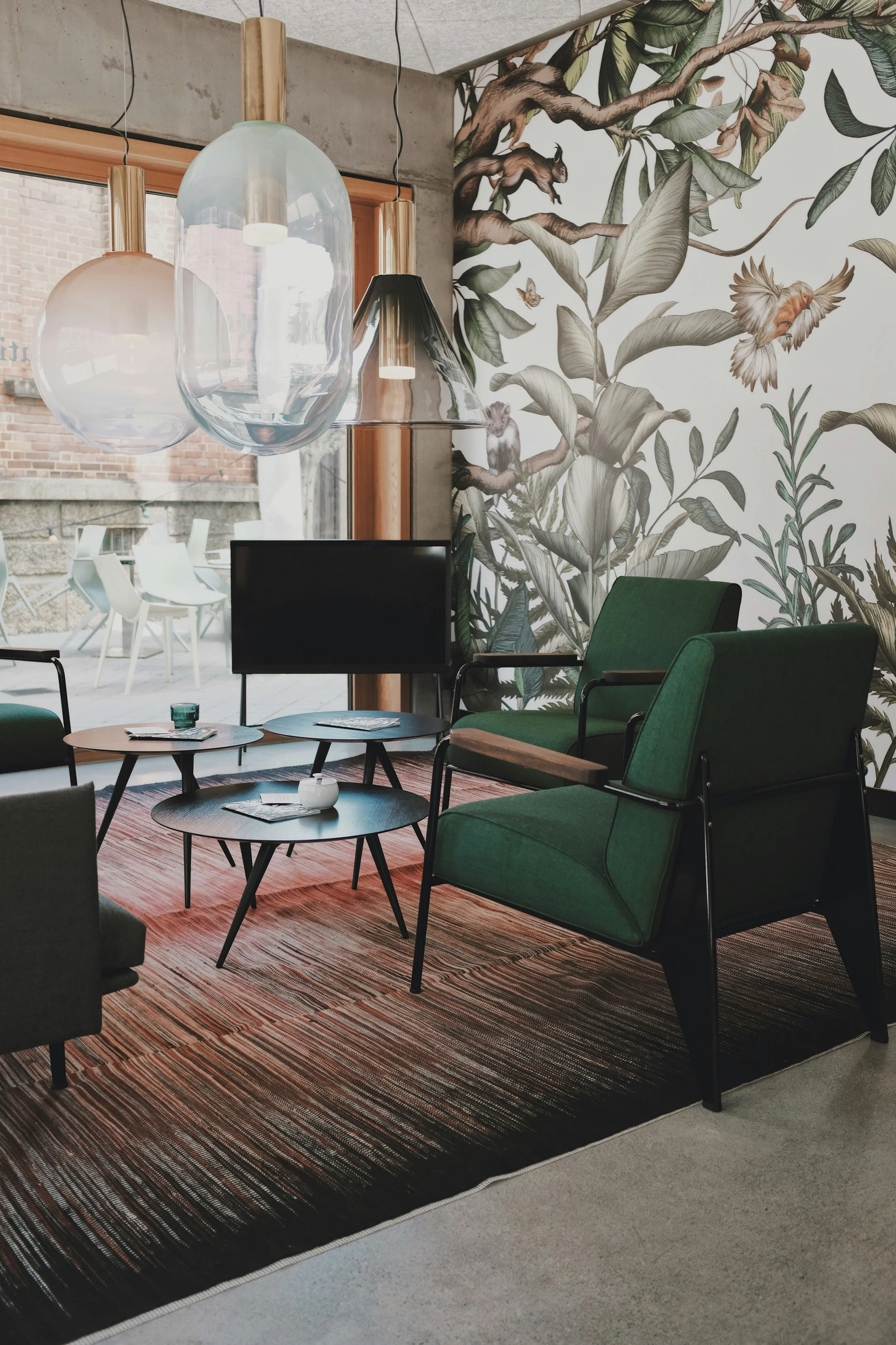 Ein moderner Aufenthaltsraum mit grünen Sesseln, einem schwarzen Couchtisch, einem grossen Wandbild mit Dschungelthema, und drei hängenden Glasschirmen über einem Couchtisch. Ein Fernseher steht in der Ecke.