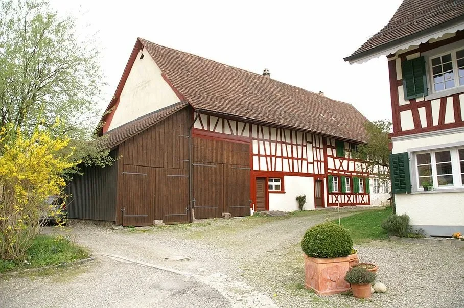 Traditionelles Fachwerkhaus mit weisser Fassade, grünen Fensterläden und rotem Holzrahmen, neben einem renovierten Scheunengebäude mit Holztoren, im Garten einige Topfpflanzen und ein gepflegter Rasen, bei sonnigem Himmel.