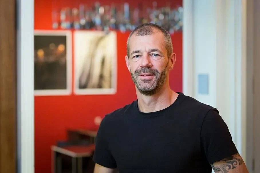 Ein Mann mit kurzem Haar und Bart trägt ein schwarzes T-Shirt, steht in einem Raum mit roter Wand und Fotografien im Hintergrund.