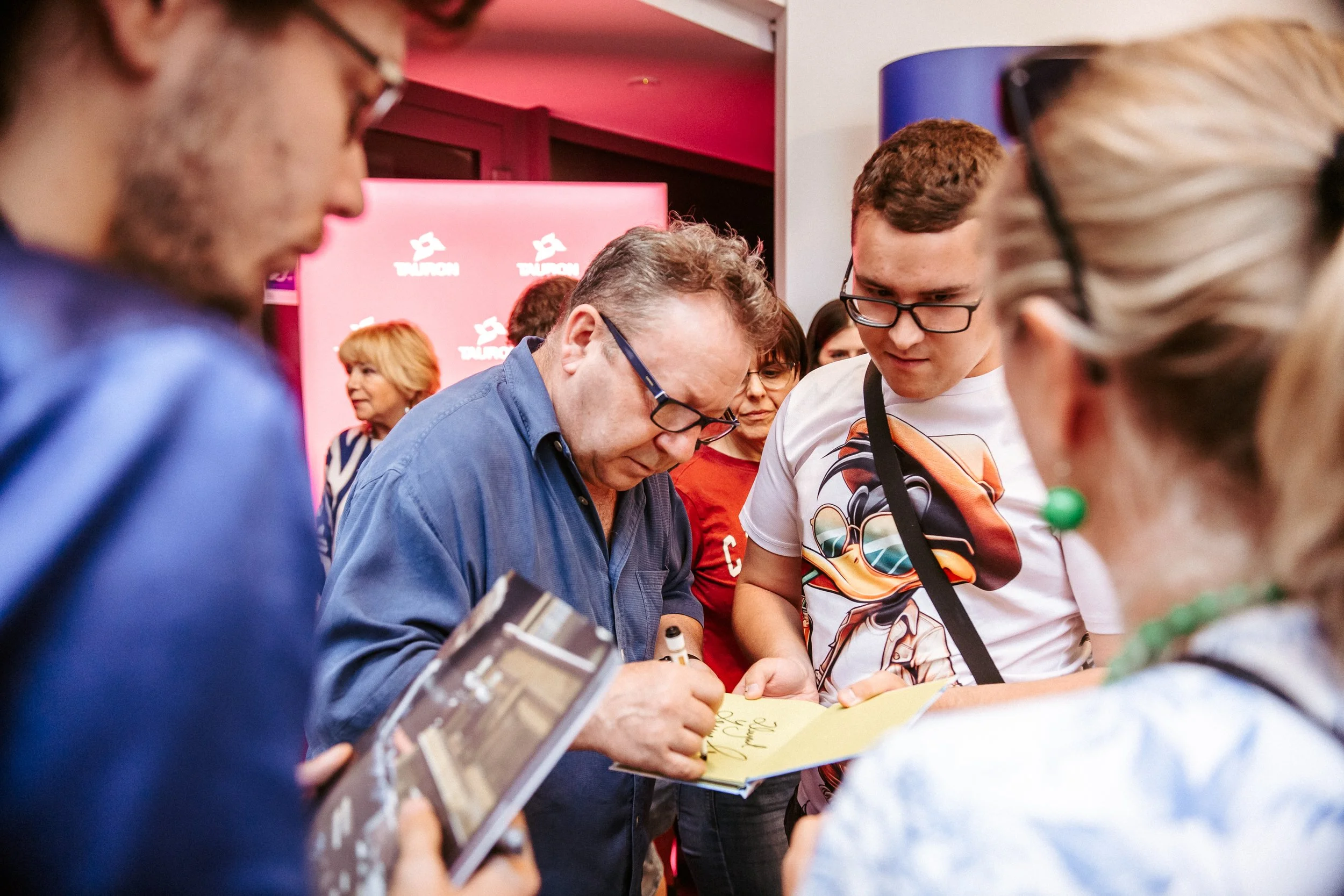 A group of people gathered around a table; one person is signing a book while others watch, with a pink Tulcom backdrop in the background.