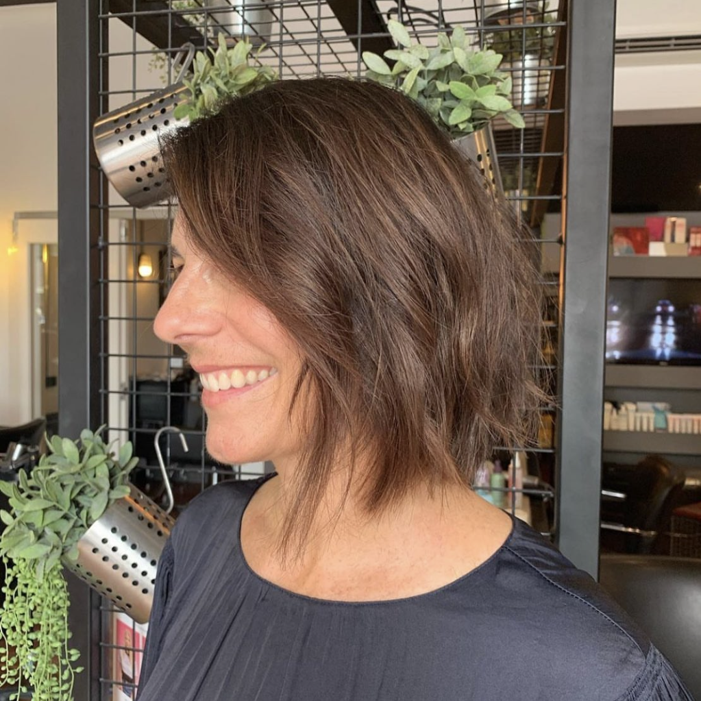 Woman with shoulder-length, wavy brown hair smiling in a modern salon setting.
