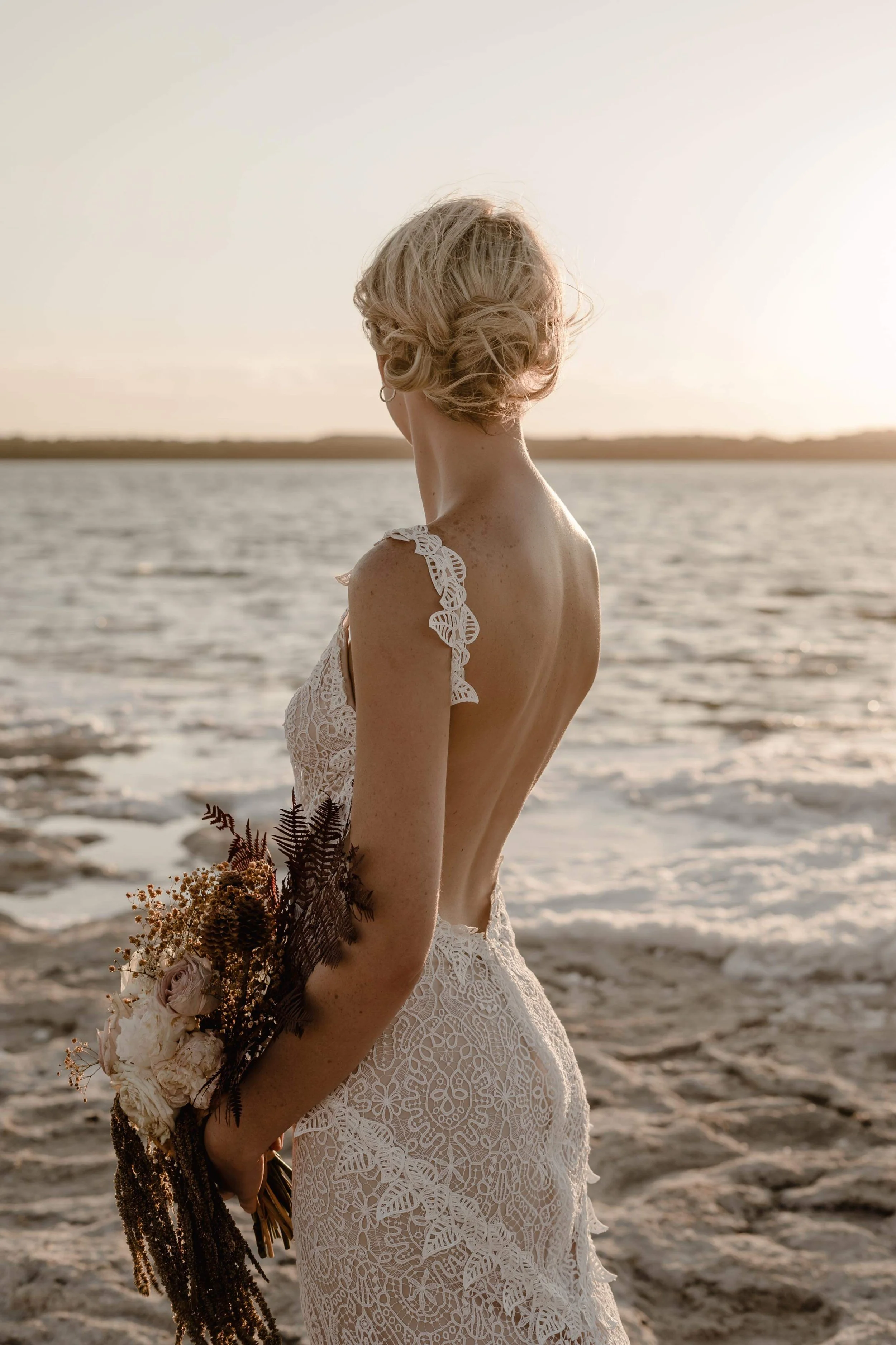 A woman in a white lace wedding dress stands by the water at sunset, holding a bouquet of flowers.