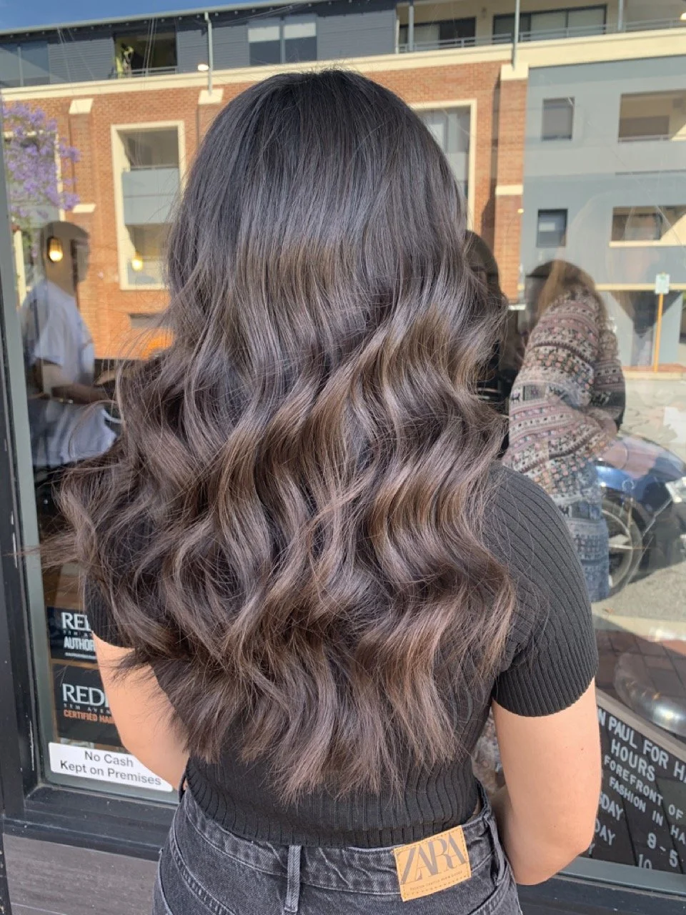 Back view of a woman with long, wavy brown hair with subtle purple highlights, wearing a black ribbed shirt and high-waisted black jeans outside a storefront.