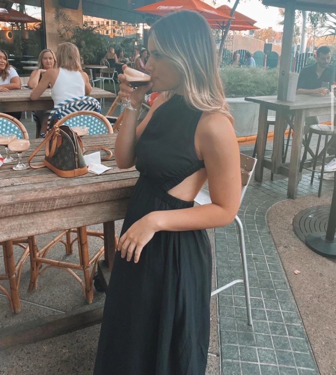A woman in a black sleeveless dress with side cutouts is drinking a cocktail at an outdoor restaurant or bar. There are other patrons seated at tables in the background, along with umbrellas and greenery.