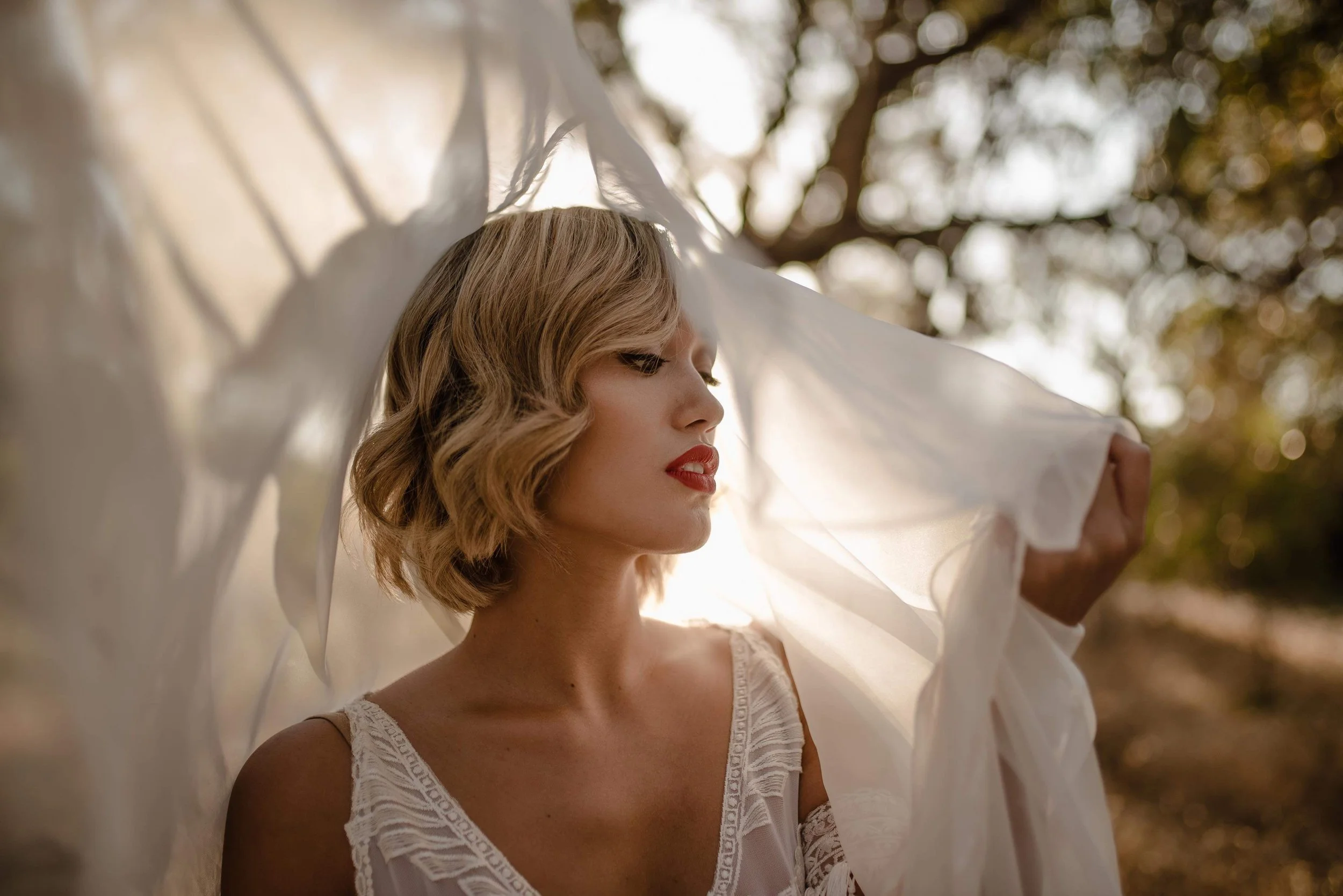 A woman with wavy blonde hair and red lipstick holds a flowing white fabric over her head outdoors with trees in the background, backlit by sunlight.