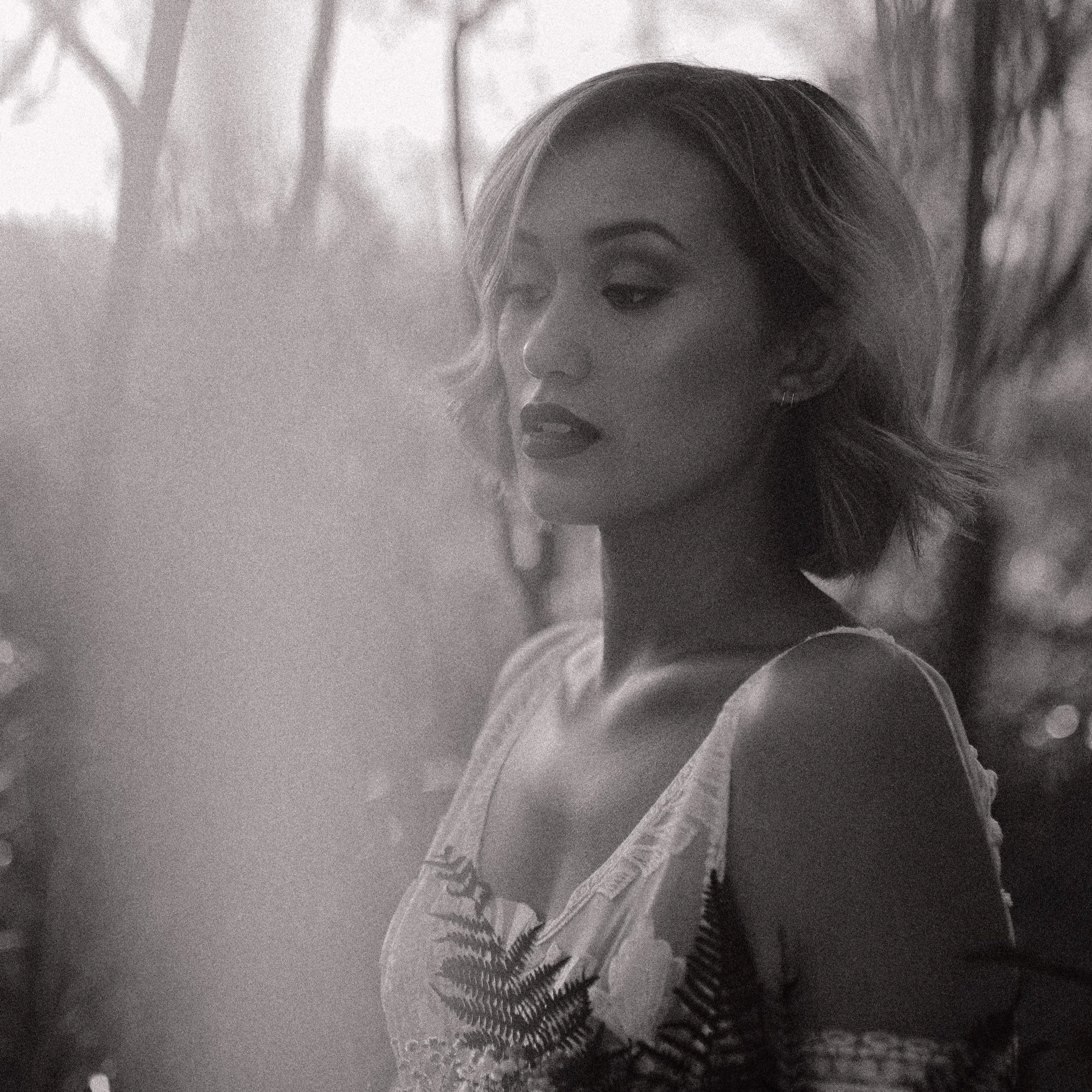 A black-and-white photo of a woman with short, wavy hair, wearing earrings and a sleeveless top with a floral pattern and fern details, standing outdoors with blurred trees in the background.