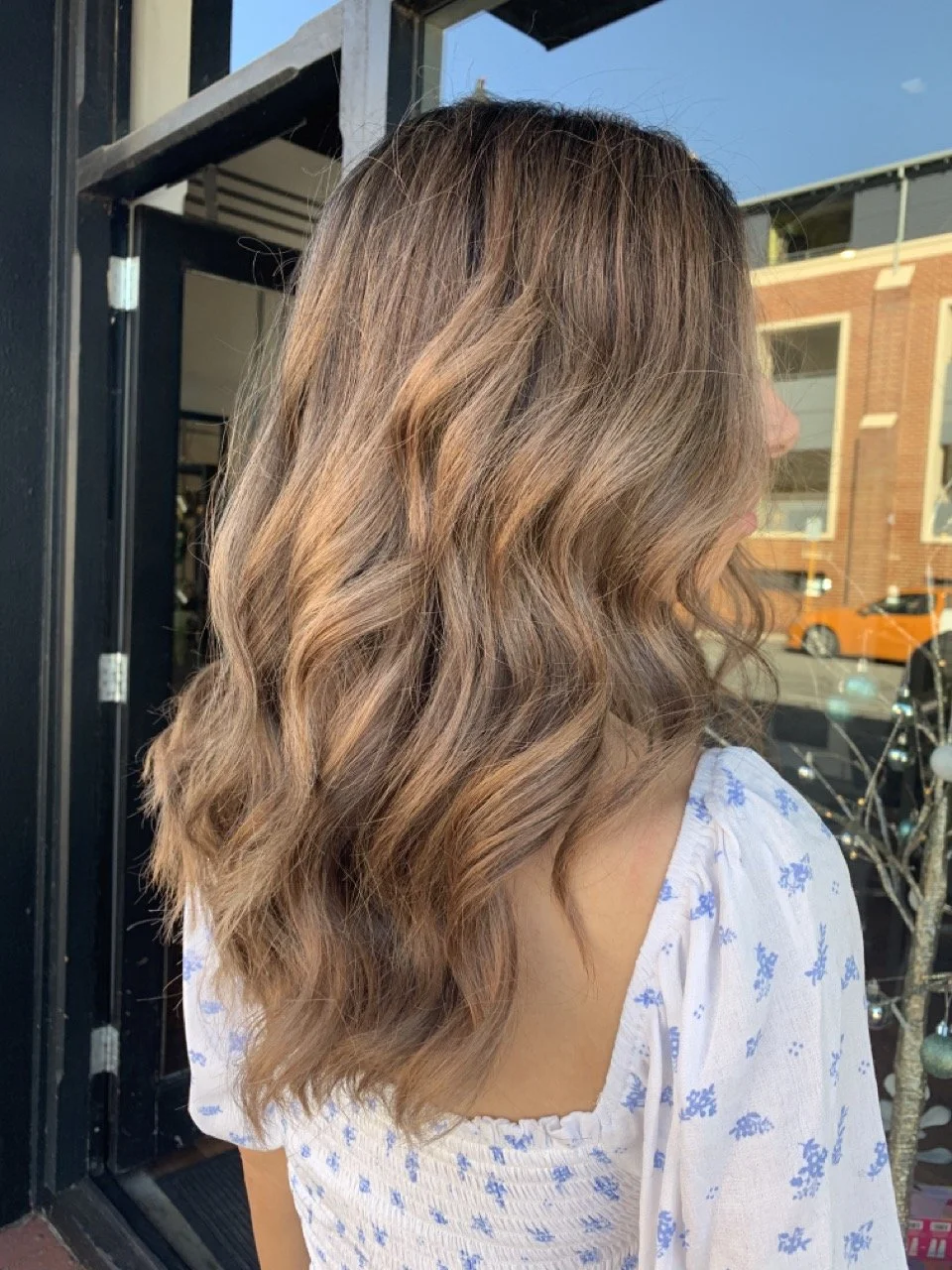 Side view of a woman with wavy, ombre brown hair standing outside near a window, wearing a white blouse with blue floral pattern.