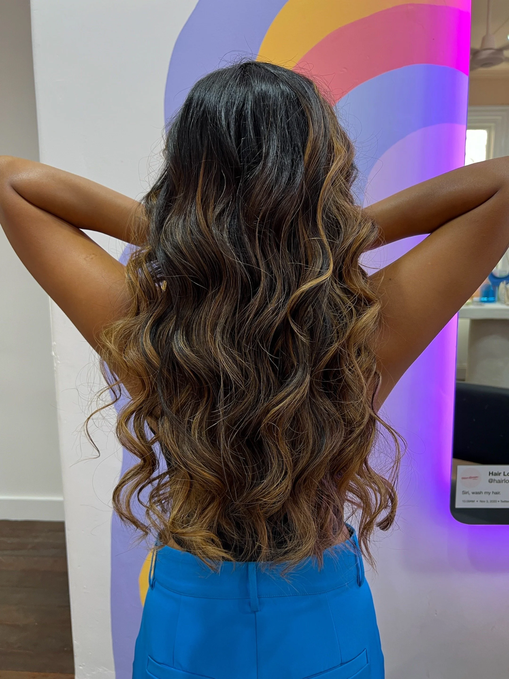 Back of a woman with long, wavy hair with highlights, wearing blue pants, standing in front of colorful artwork.