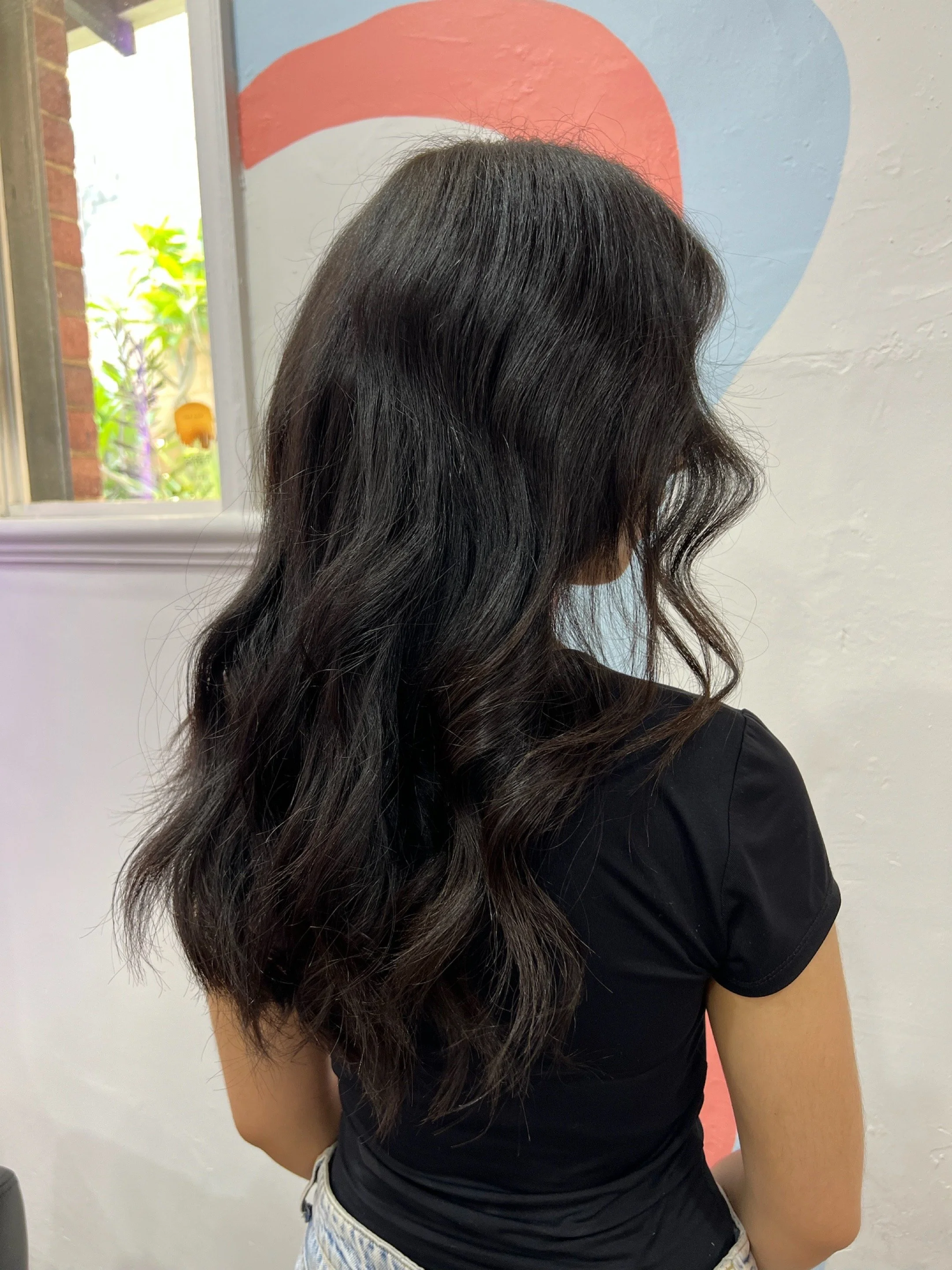 A woman with dark, wavy hair standing indoors near a white wall with a painted design, facing away from the camera.