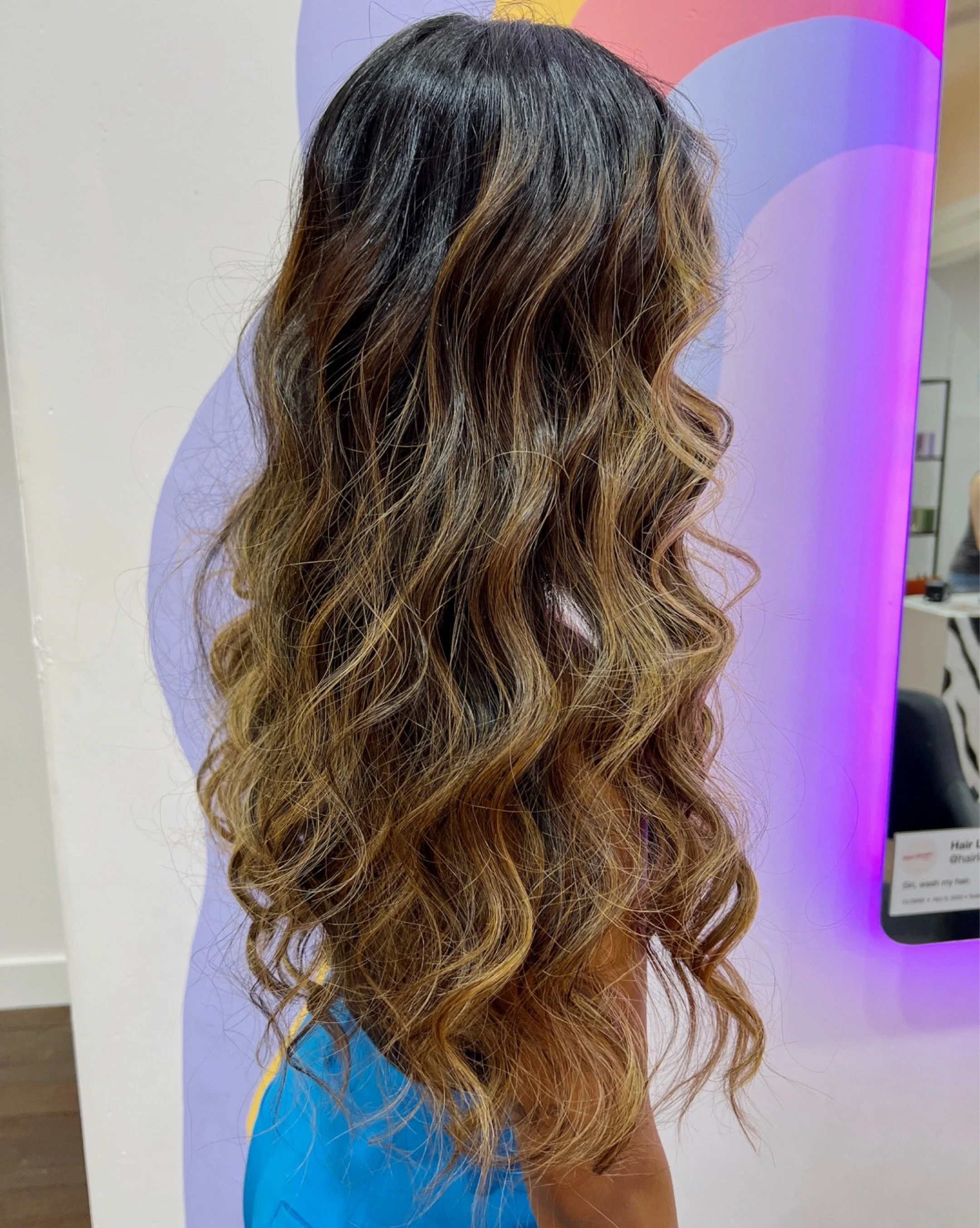 Side view of a woman with long, wavy, ombre hair that transitions from dark at the roots to light at the ends, standing in front of a pastel-colored wall.