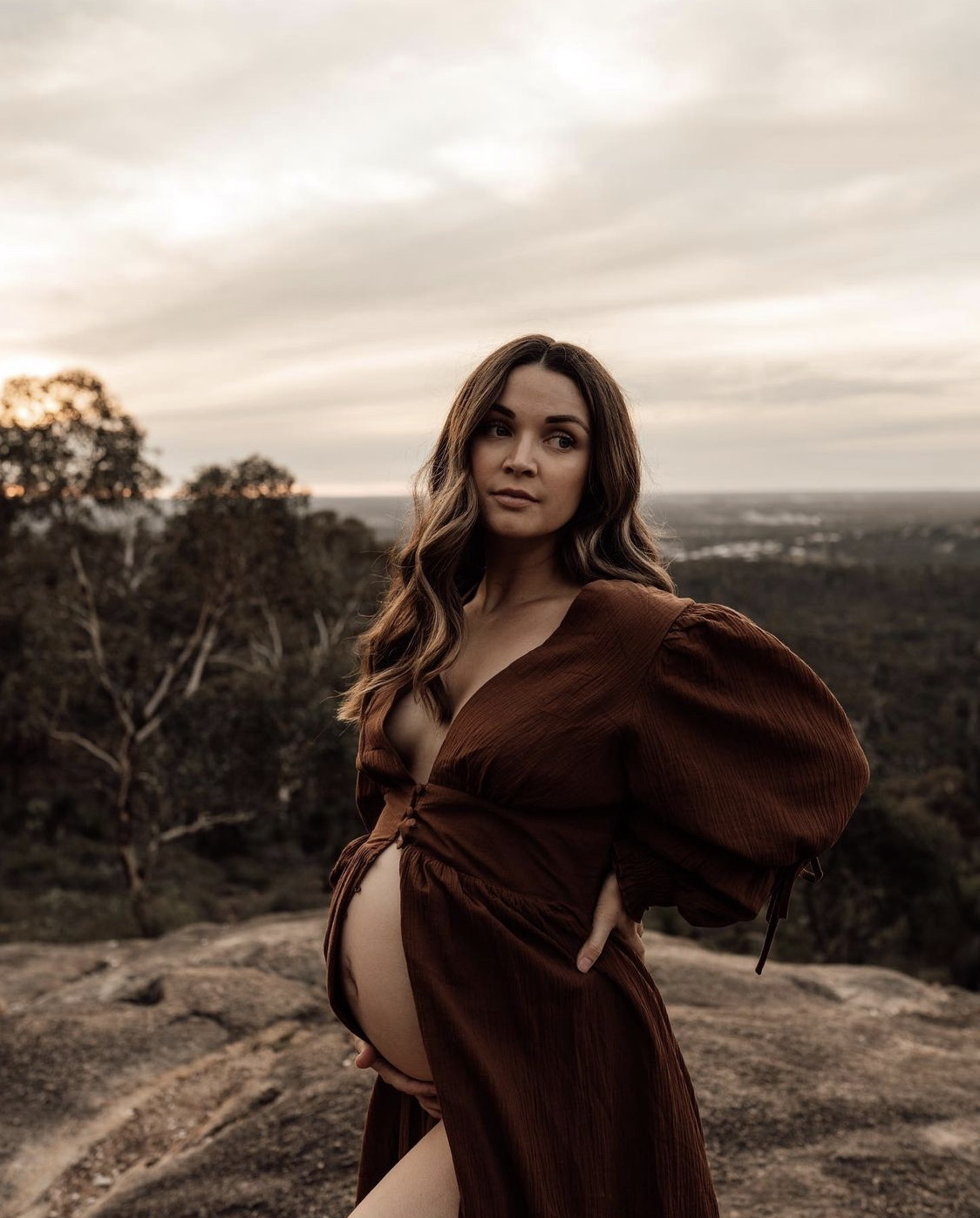 A pregnant woman in a brown dress standing outdoors on a rocky landscape with trees and a cloudy sky in the background.
