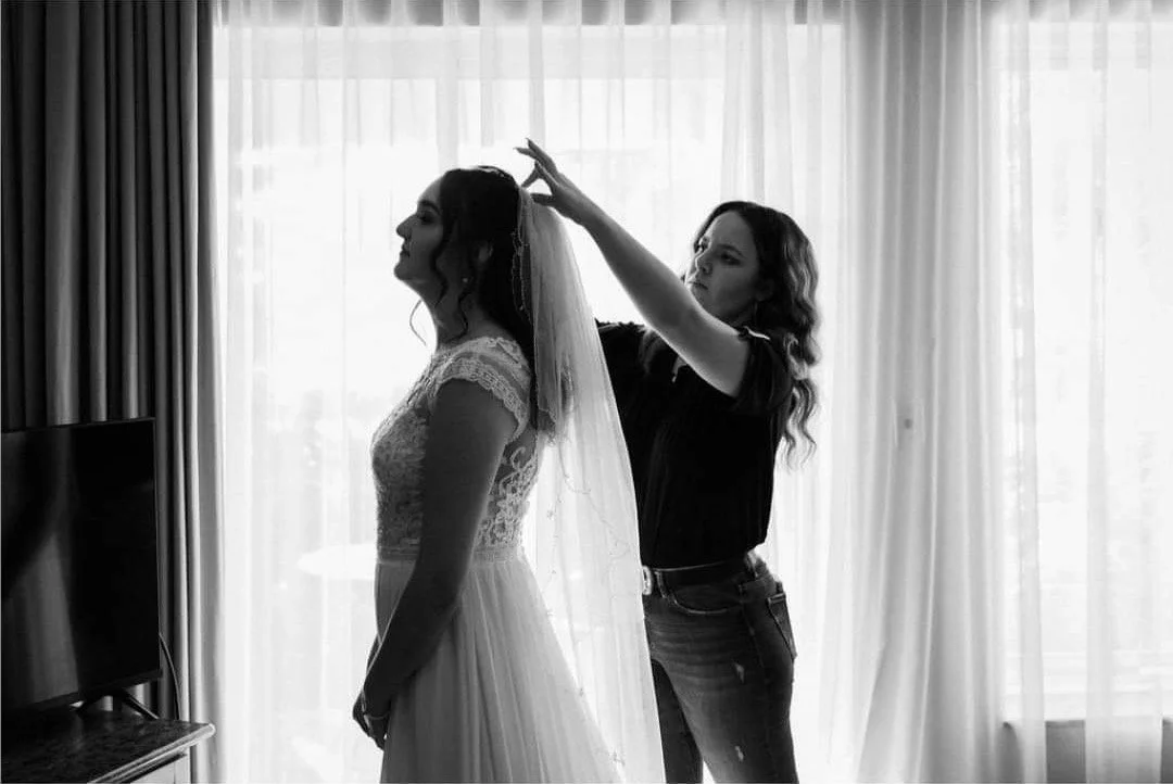 A woman in a lace wedding dress stands with eyes closed as another woman adjusts her veil in a bright room with sheer curtains.