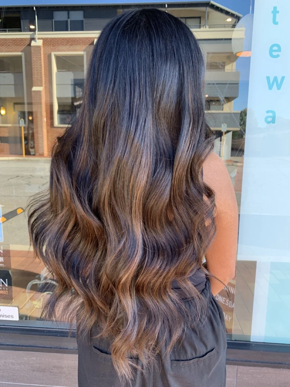 Back view of a woman with long, wavy hair with dark roots and caramel-brown highlights standing outside near a window.