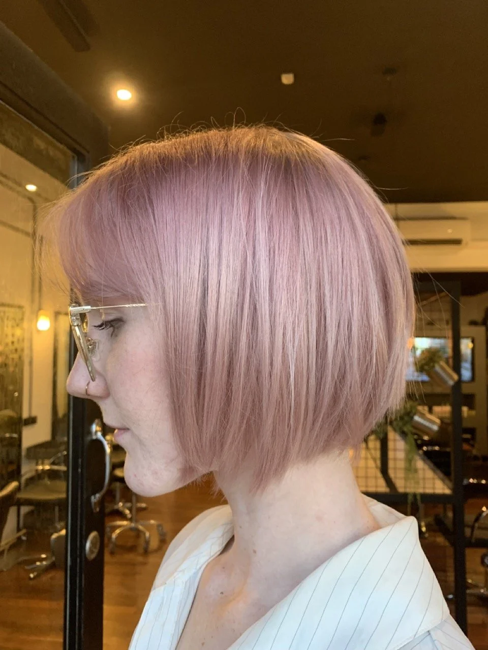 A woman with short, pinkish-blonde hair in a modern salon, wearing glasses and a white striped shirt, standing in profile.