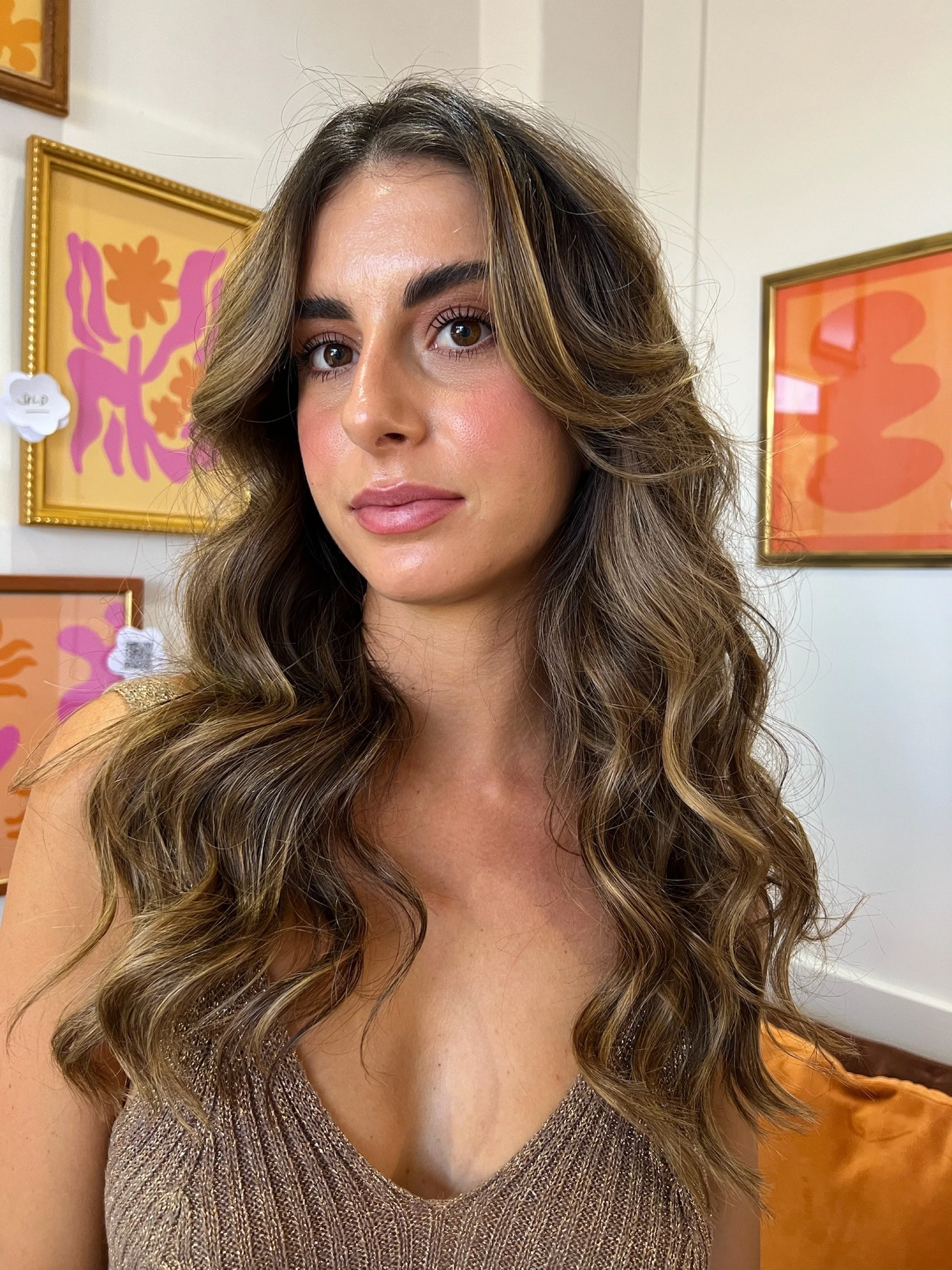 A young woman with long, wavy brown hair and light makeup, wearing a shimmery beige top, standing in front of colorful abstract artwork.