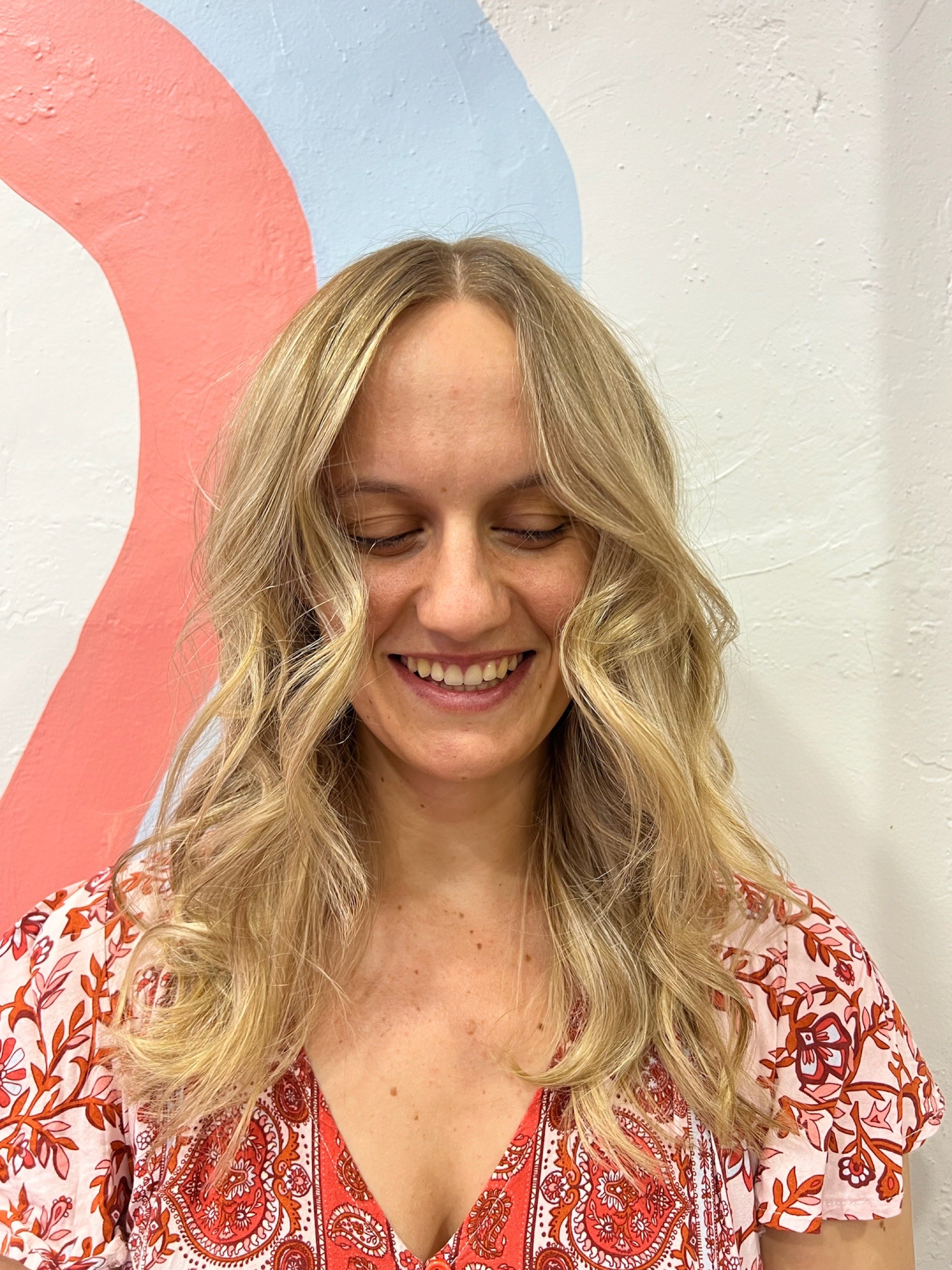 Woman with wavy blonde hair smiling with eyes closed against a wall with pink and blue design.