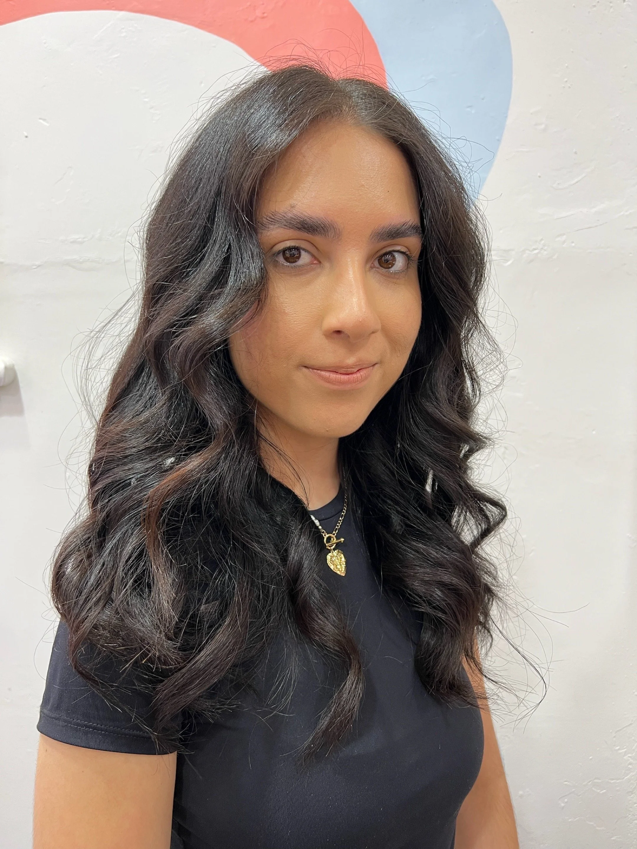 A woman with long, wavy dark hair, wearing a black t-shirt and a gold necklace, standing in front of a white wall with a small, partially visible painted design.