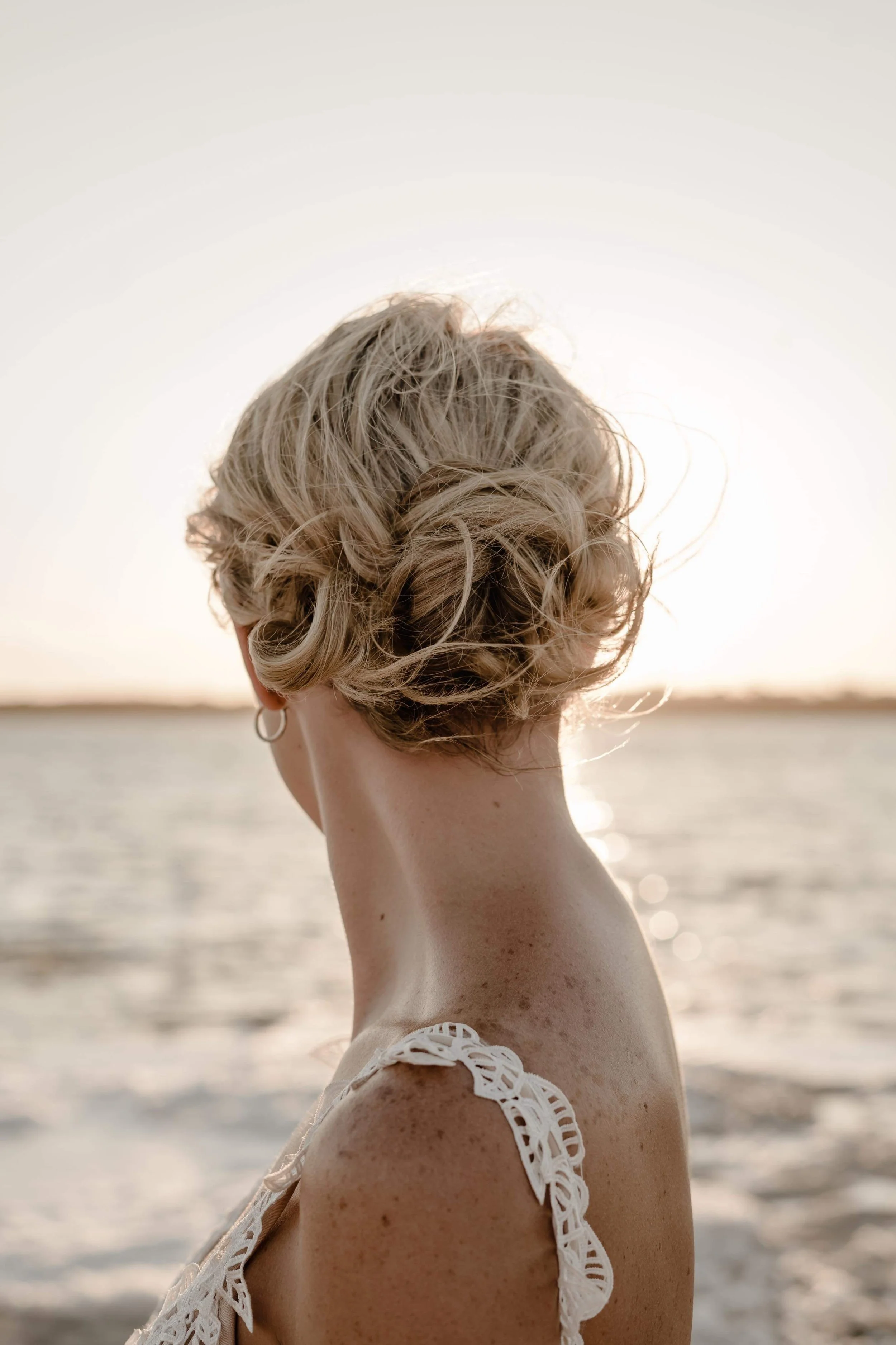 A woman with short, curly blonde hair looking away near a body of water at sunset.
