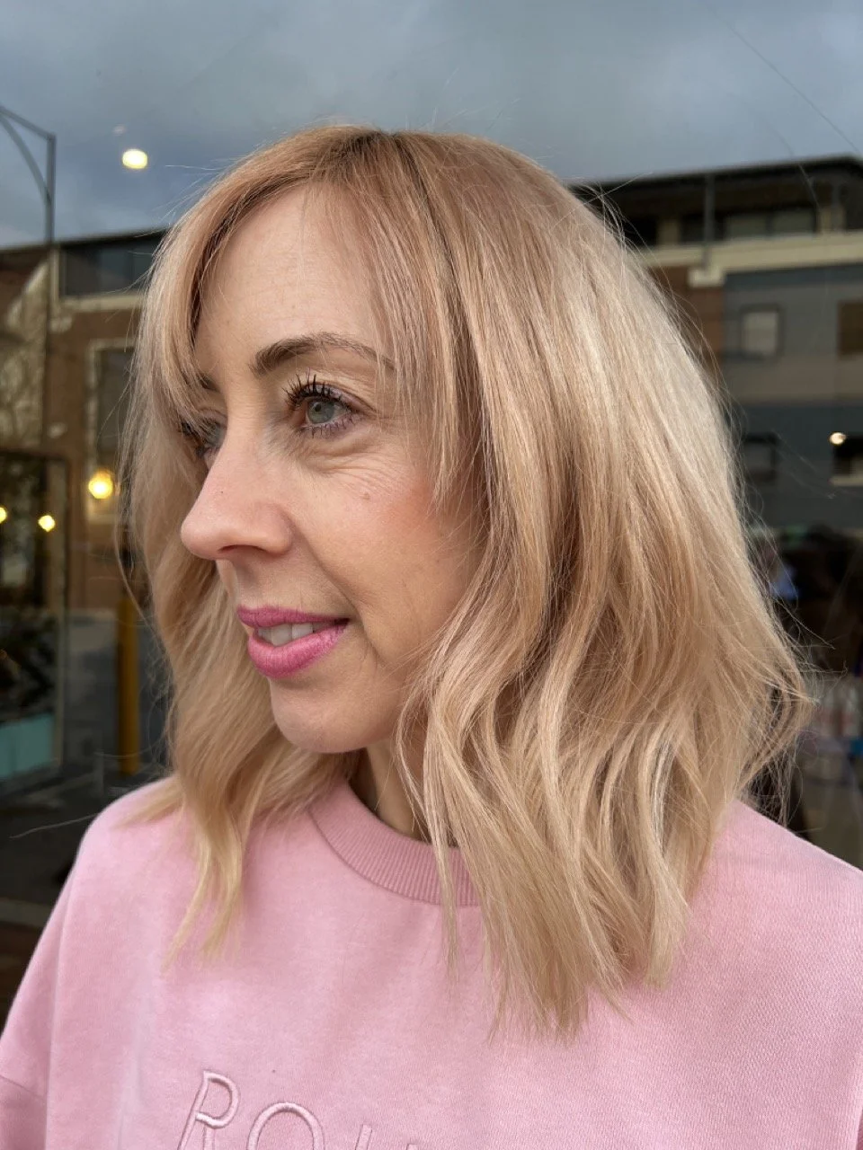 Side profile of a woman with shoulder-length blonde hair and blue eyes, wearing a light pink sweatshirt, standing outdoors in front of a building with large windows and warm lighting.