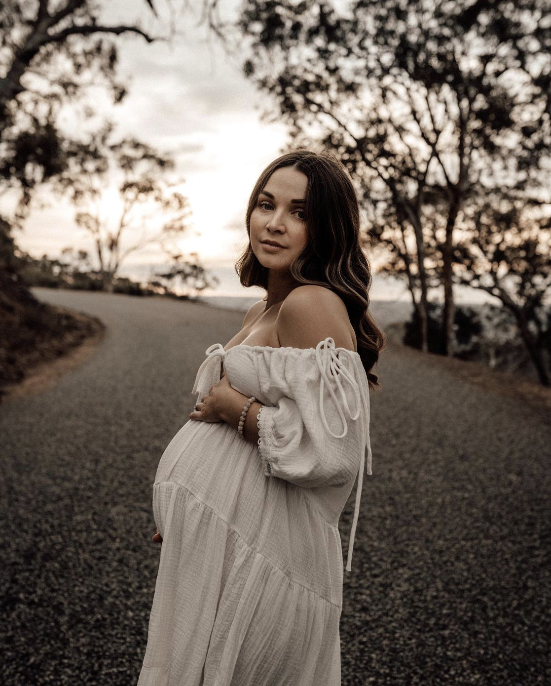 A pregnant woman in a white dress stands on a winding gravel road during sunset, with trees in the background.