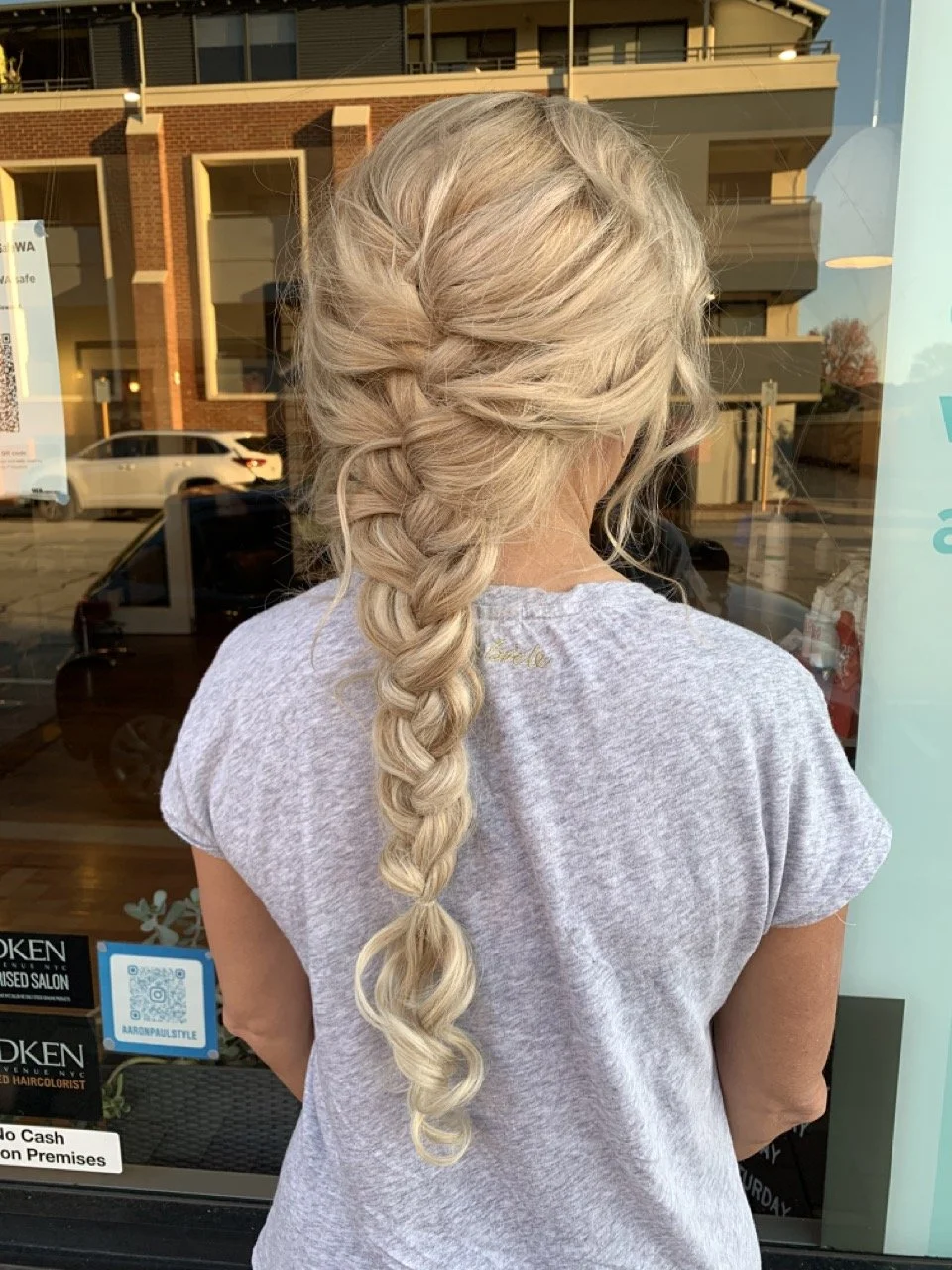 Back view of a woman with long blonde hair styled in a loose, tousled braid, wearing a gray casual t-shirt, standing outside a salon window.