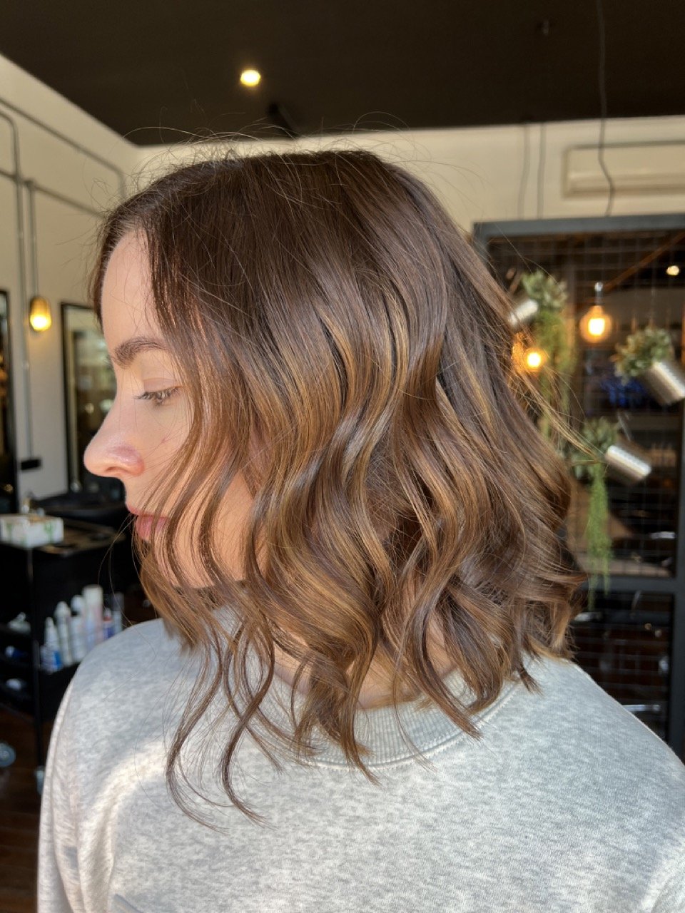 Side view of a woman with medium-length, wavy, brown hair with lighter highlights, in a salon with warm lighting and decorative plants.