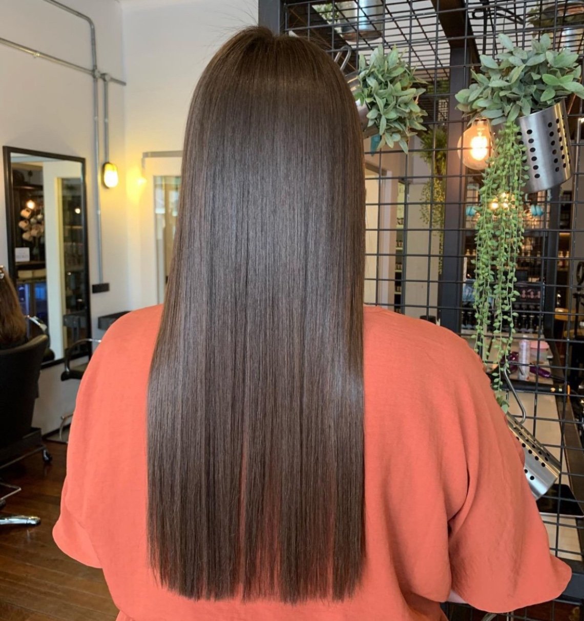 Back view of a woman with long, straight, shiny brown hair in a salon. She is wearing an orange top, and there are green plants and salon mirrors in the background.