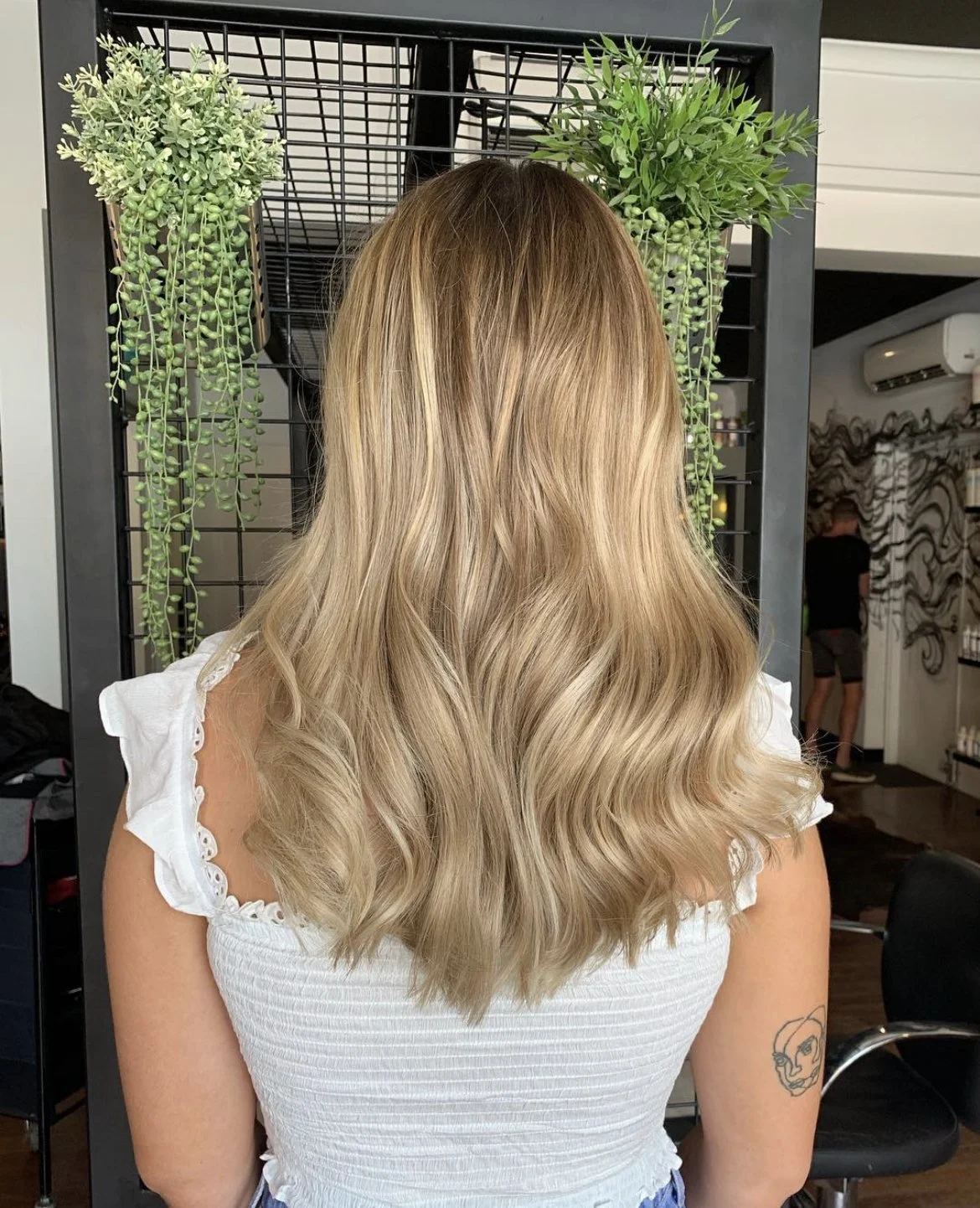 Back view of a woman with long, wavy blonde hair sitting in a salon, with green hanging plants and salon equipment in the background.