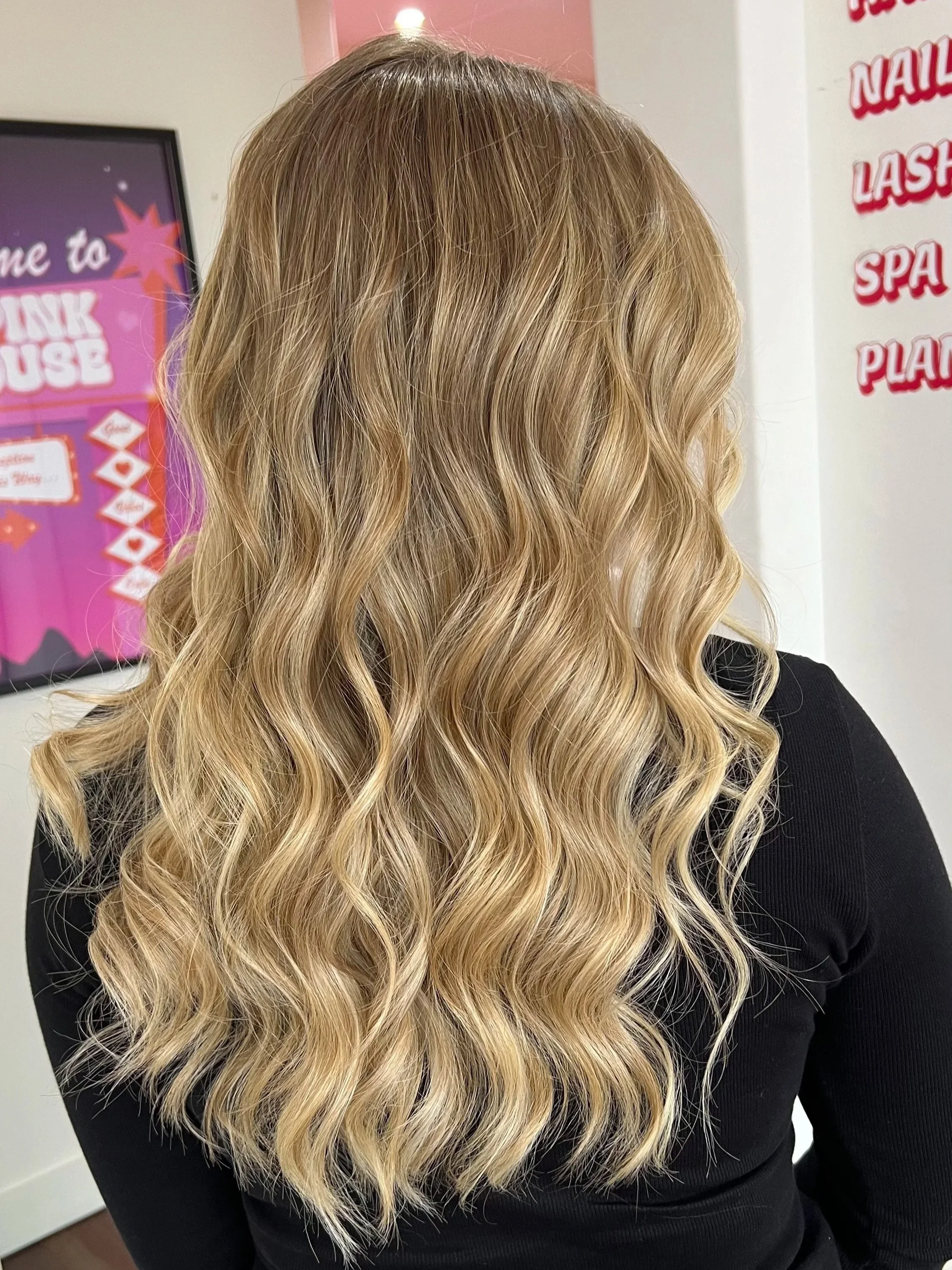 Back view of a woman with long, wavy blonde hair wearing a black top, inside a salon with pink and white decor.