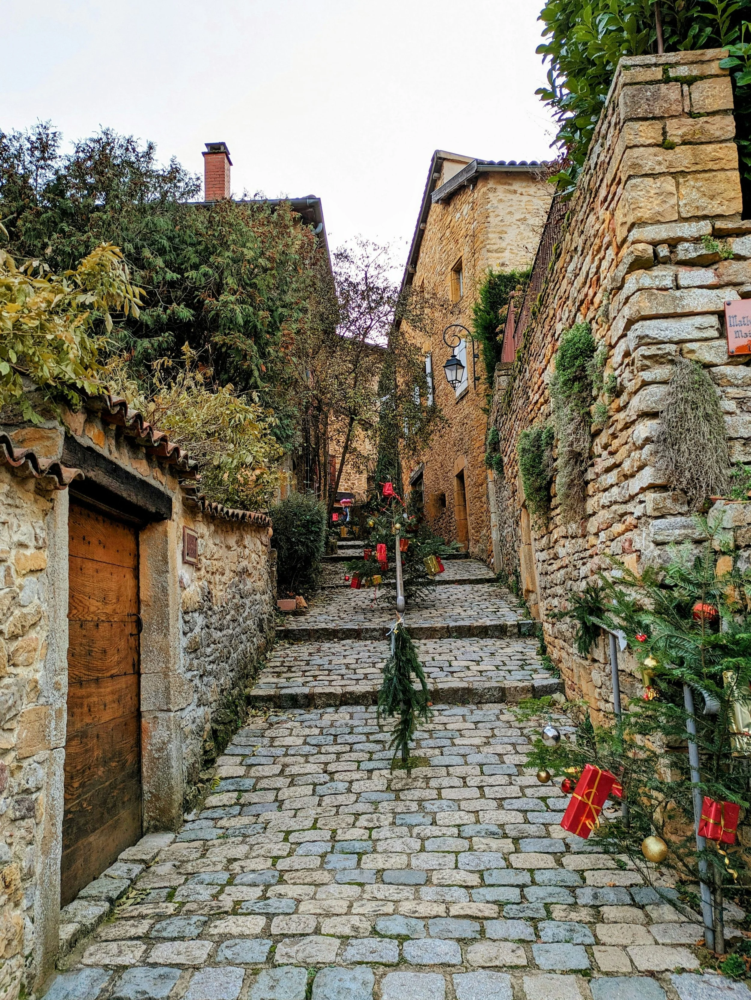 Cobblestone street in a European village with Christmas decorations and a small Christmas tree. The street is lined with stone buildings and greenery, with a lamp post and steps leading uphill.