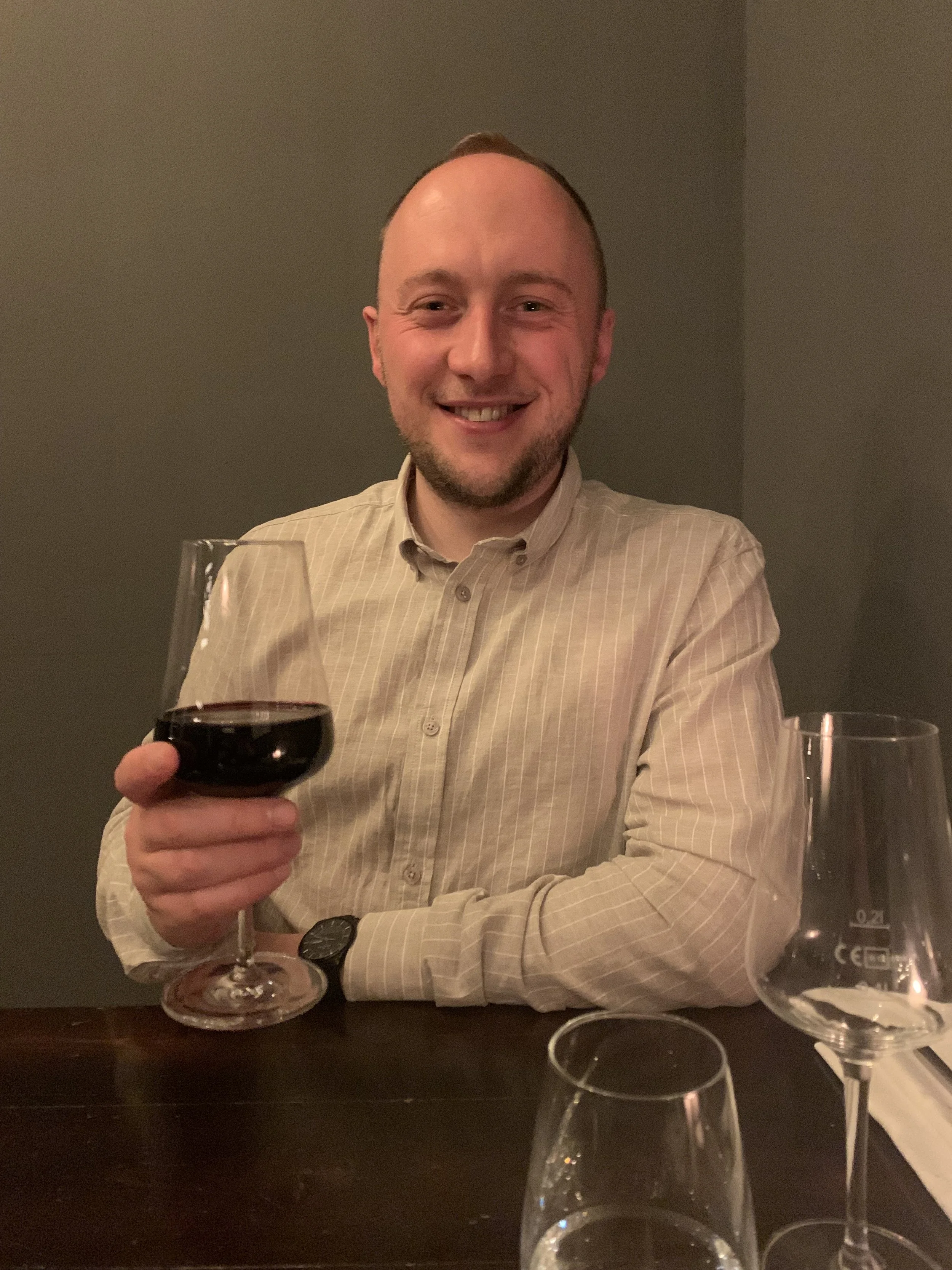 A smiling man in a beige striped shirt sitting at a table holding a glass of red wine in his right hand, with empty wine glasses on the table.