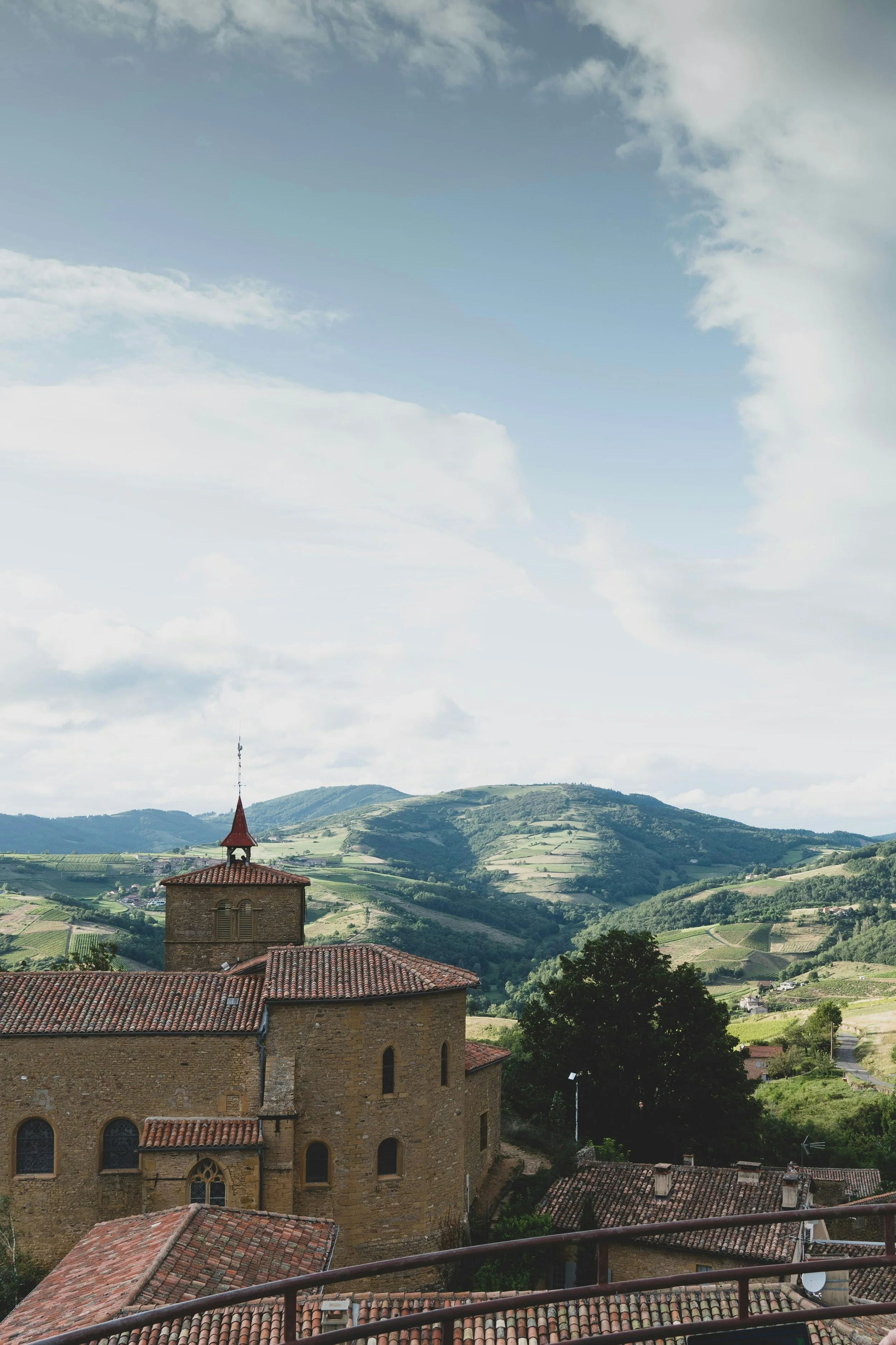 A scenic view of a historical stone church with a red-tiled roof and a small bell tower, set in a lush green hilly landscape under a partly cloudy sky.