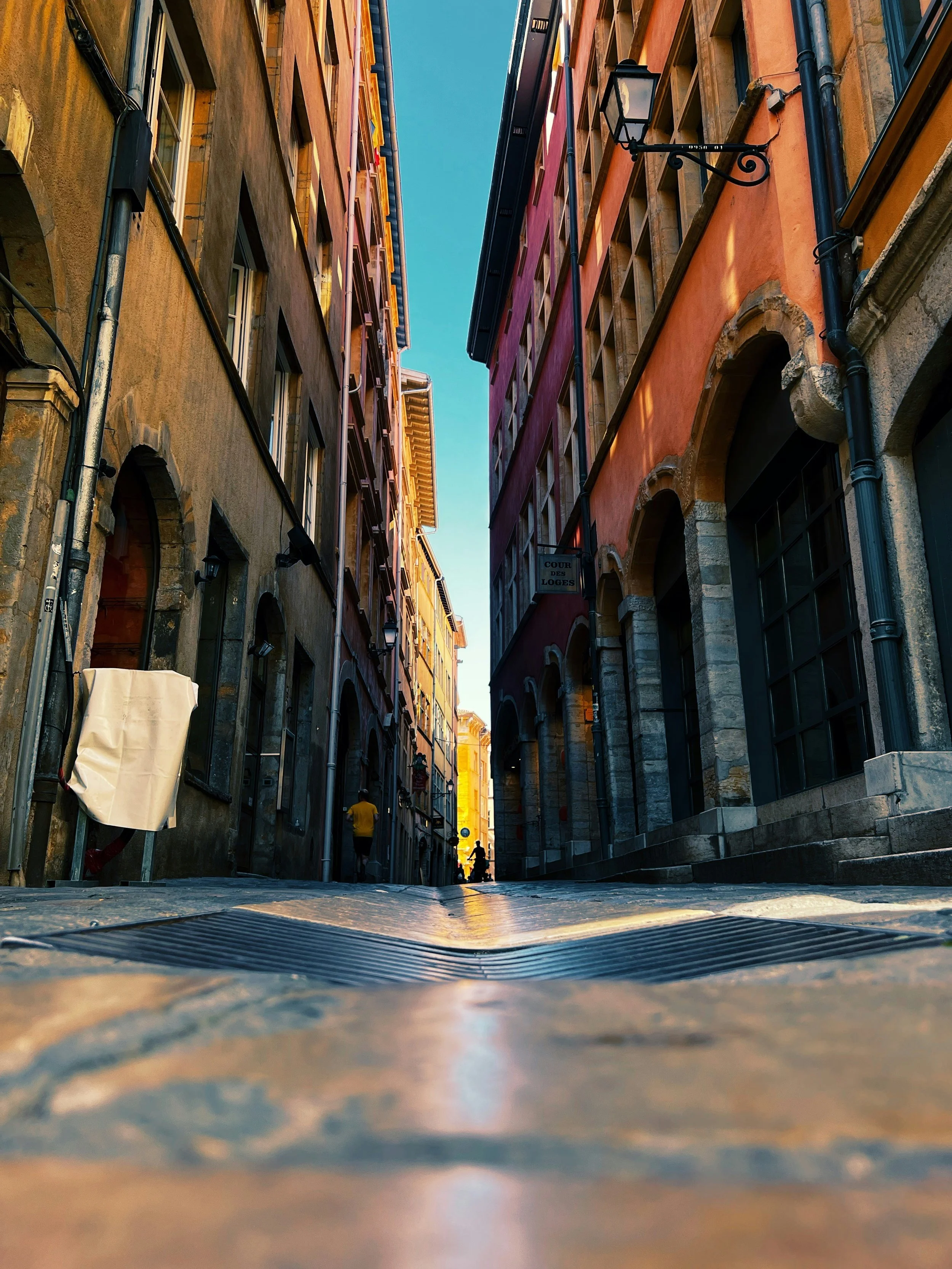 A narrow European street with colorful buildings, arched doorways, and cobblestone pavement, with a few pedestrians walking in the distance under a clear blue sky.