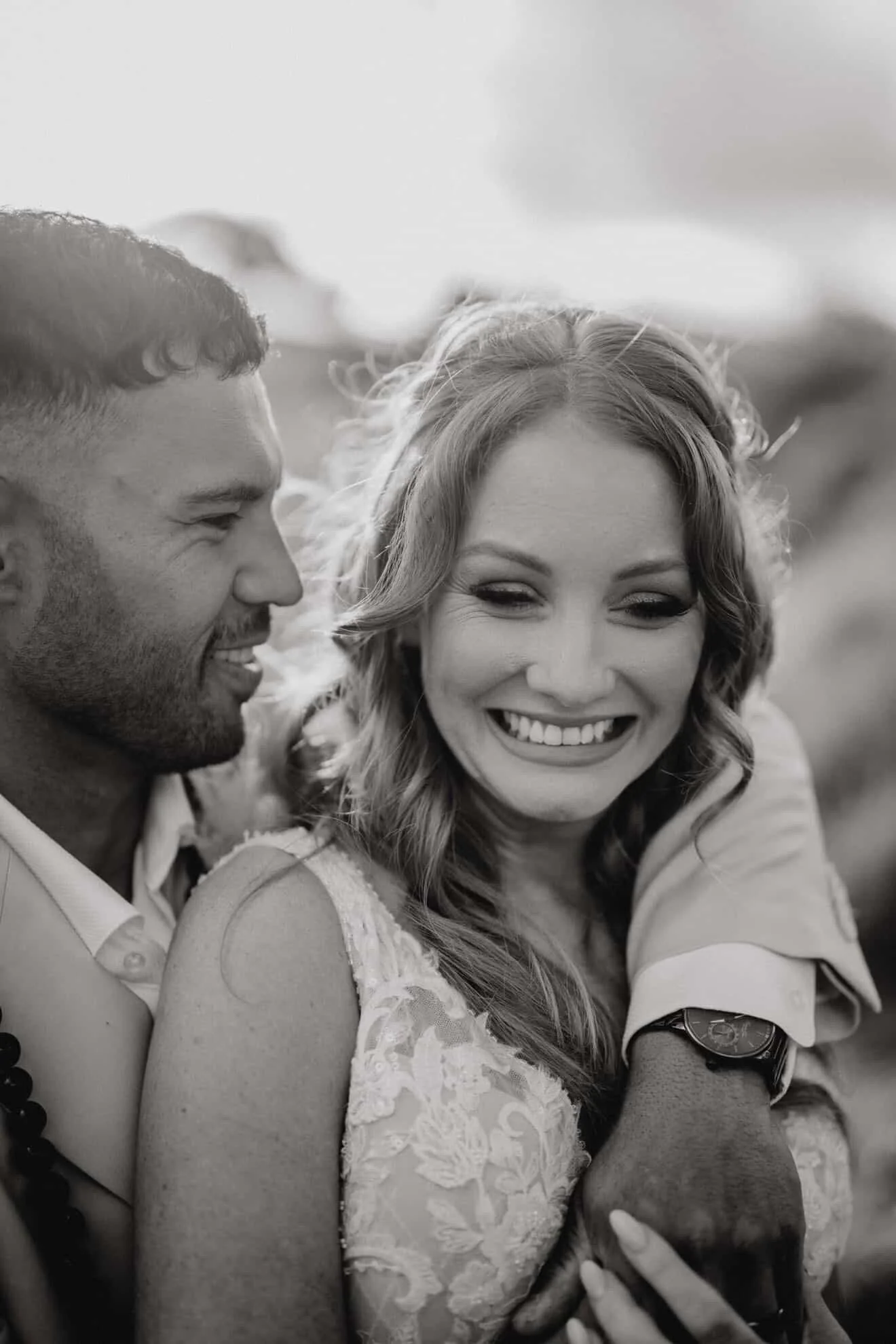 A smiling man and woman taking a selfie together outdoors. The woman has wavy hair and is wearing a lace top, while the man has short curly hair and is dressed casually.