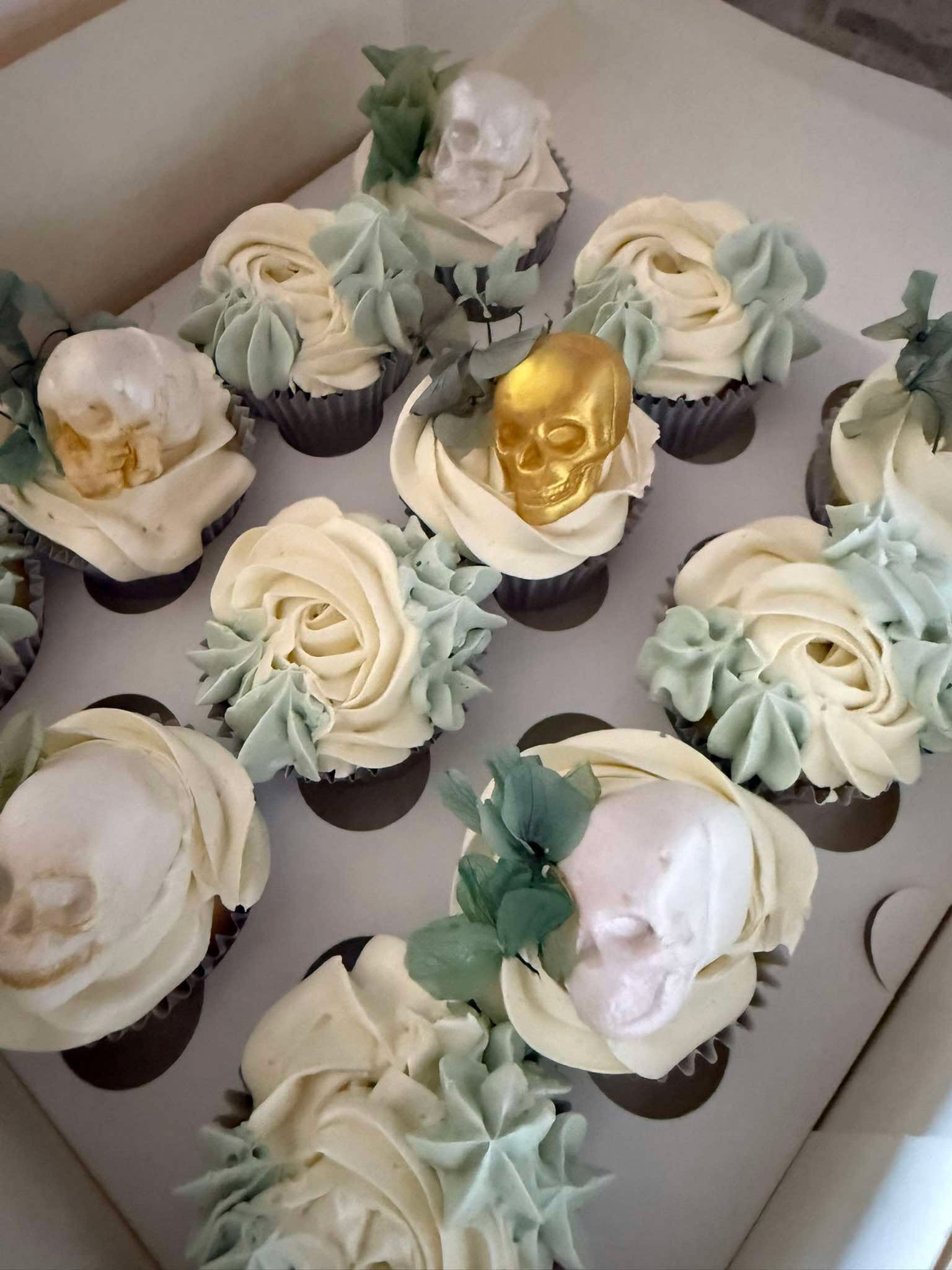 Box of cupcakes decorated with white and light green frosting and skull toppers, some painted gold.