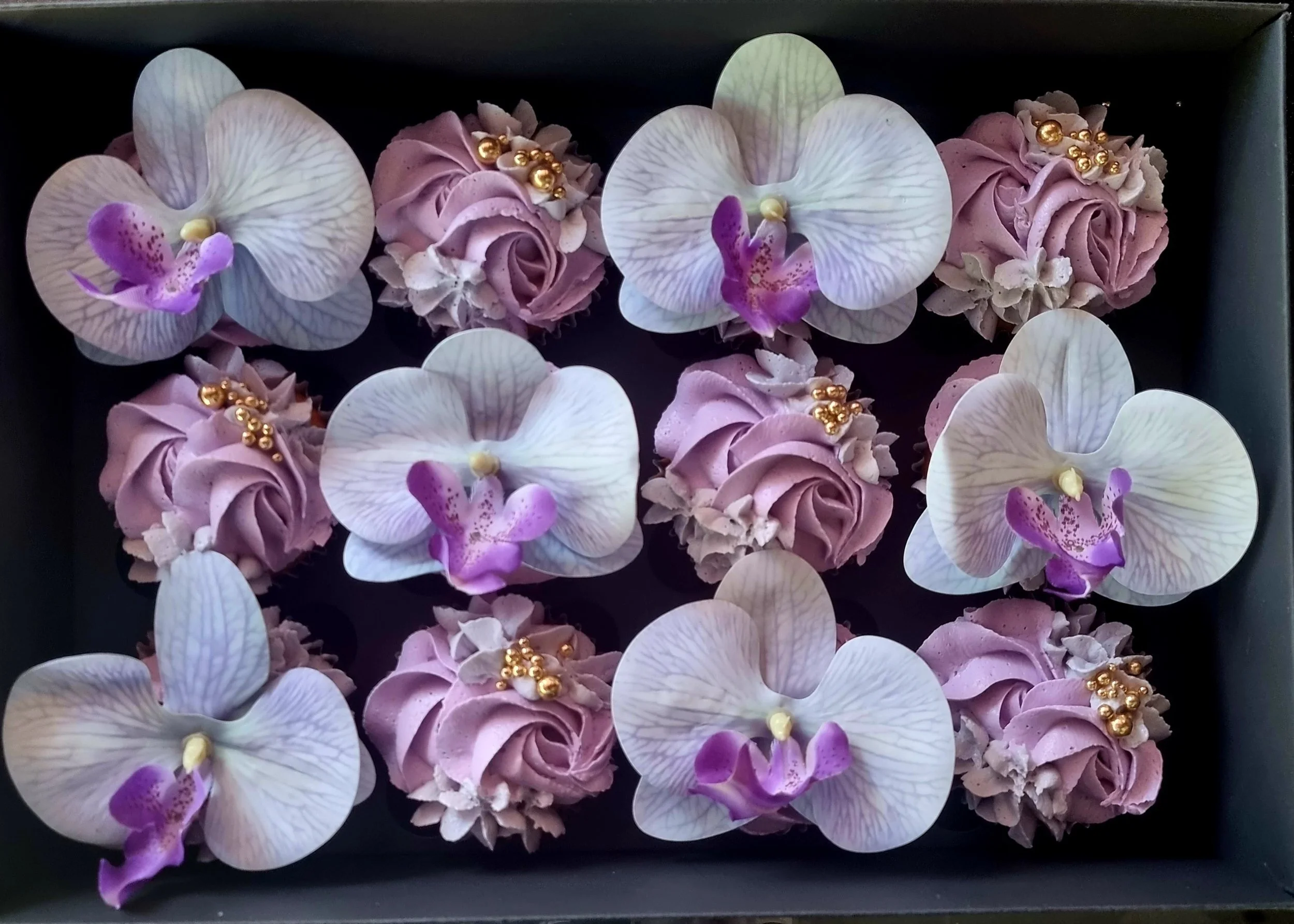 Box of decorated cupcakes topped with white orchid and purple flower edible decorations, with gold bead accents.
