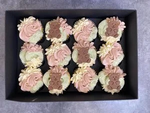 A black tray holding twelve decorated cupcakes with pink and beige frosting, some adorned with bear-shaped fondant decorations.