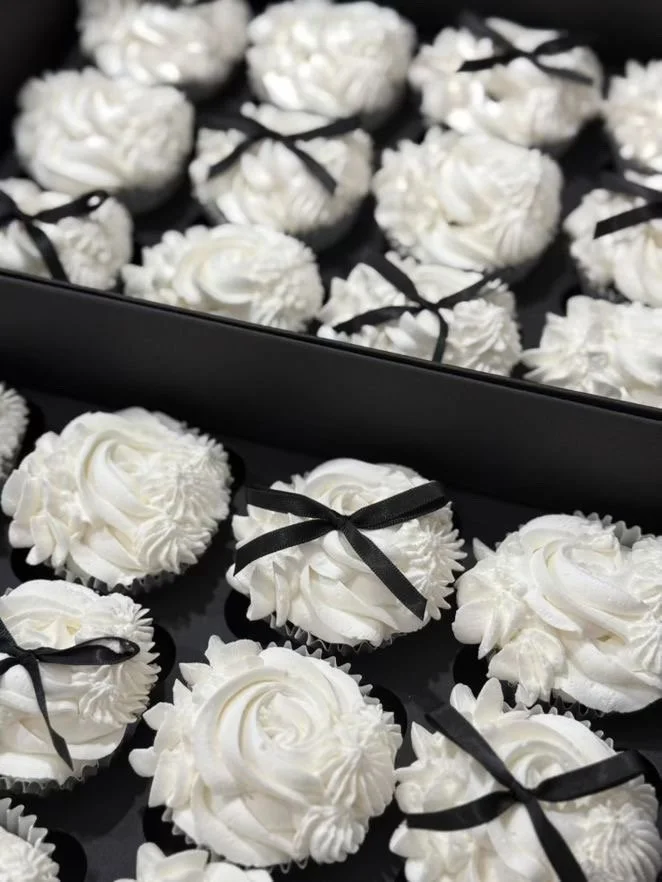 Tray of white cupcakes decorated with piped frosting and small black ribbons.