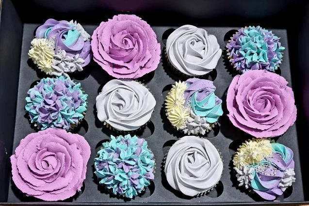 A tray of decorated cupcakes with pastel-colored frosting in floral and rosette designs, featuring purple, white, light blue, and yellow colors.
