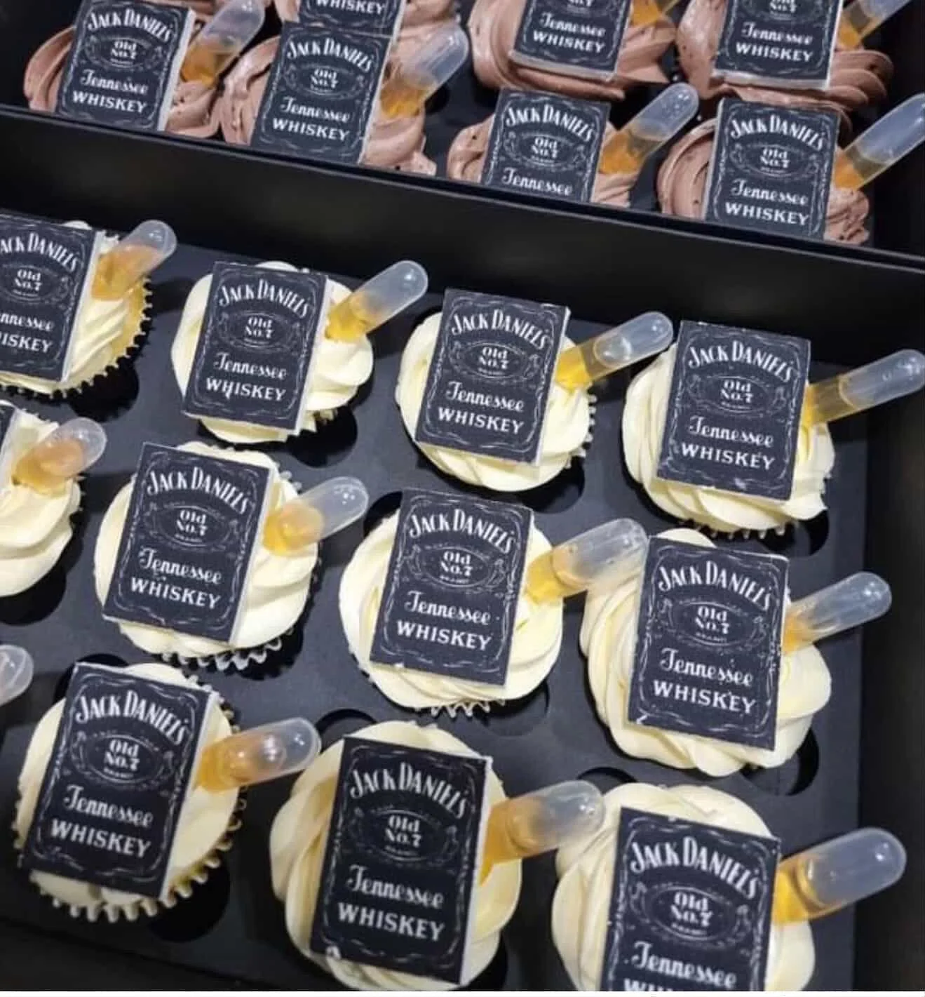 Cupcakes decorated with Jack Daniel's Tennessee Whiskey labels and small syringe-like toppers filled with whiskey on a black tray.