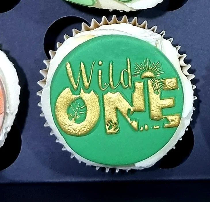 Cupcake with green frosting and gold lettering that says 'Wild ONE' with a small leaf, a sun, and a tree design.
