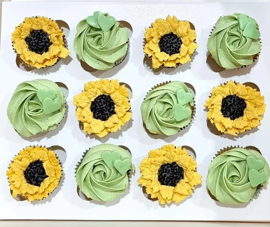 Assorted cupcakes arranged in a white box, featuring green and yellow frosting with black and green decorative toppings.