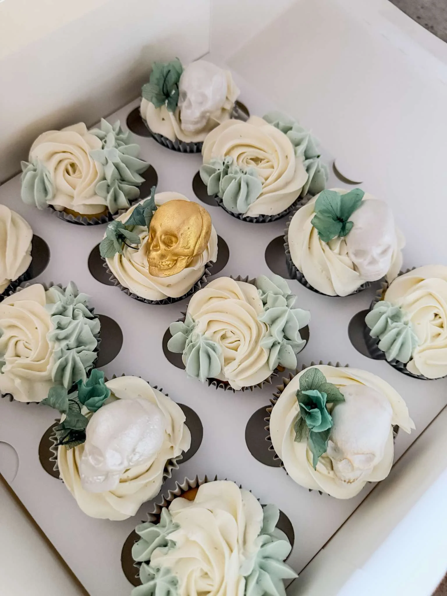 A box of cupcakes decorated with white and light green frosting, some featuring a skull and floral designs.