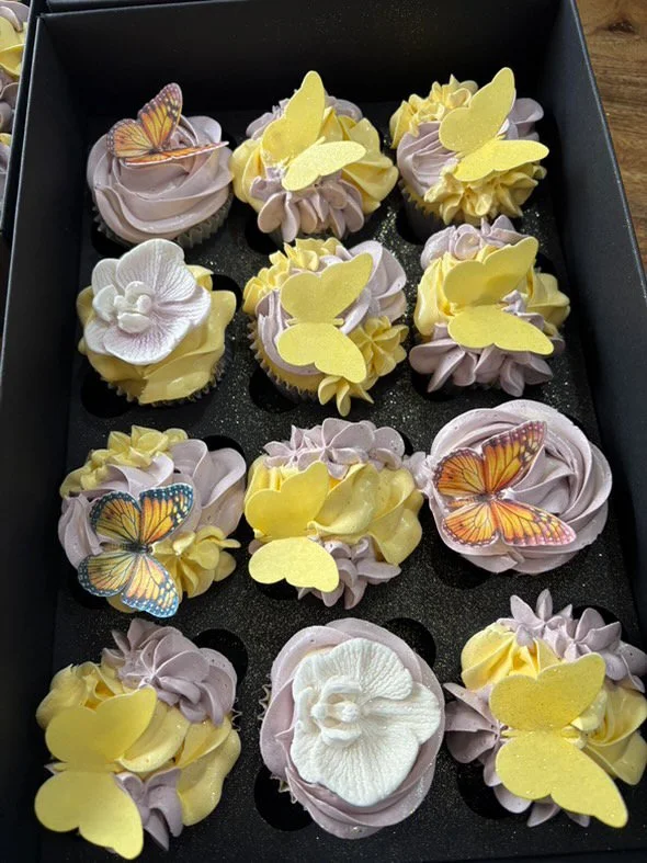 Assorted decorated cupcakes with yellow, purple, and white frosting, topped with butterfly and heart-shaped toppers inside a black box.