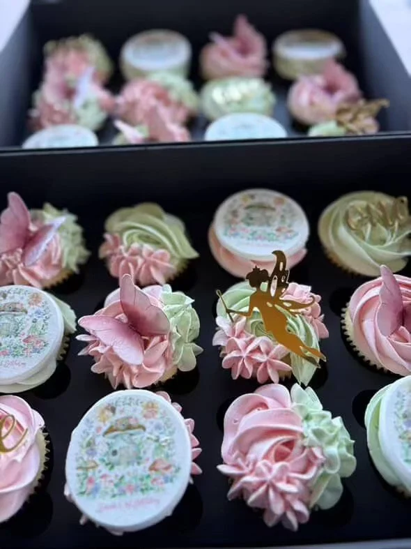 Assorted decorated cupcakes with pastel-colored frosting, some with pink and green buttercream and others with edible images, arranged in a black box. Some cupcakes have small decorative gold and pink accents.