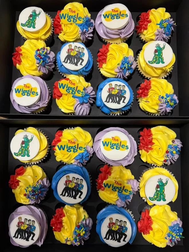 Box of cupcakes decorated with colors and toppers themed from the children's show 'The Wiggles,' including characters and logos.